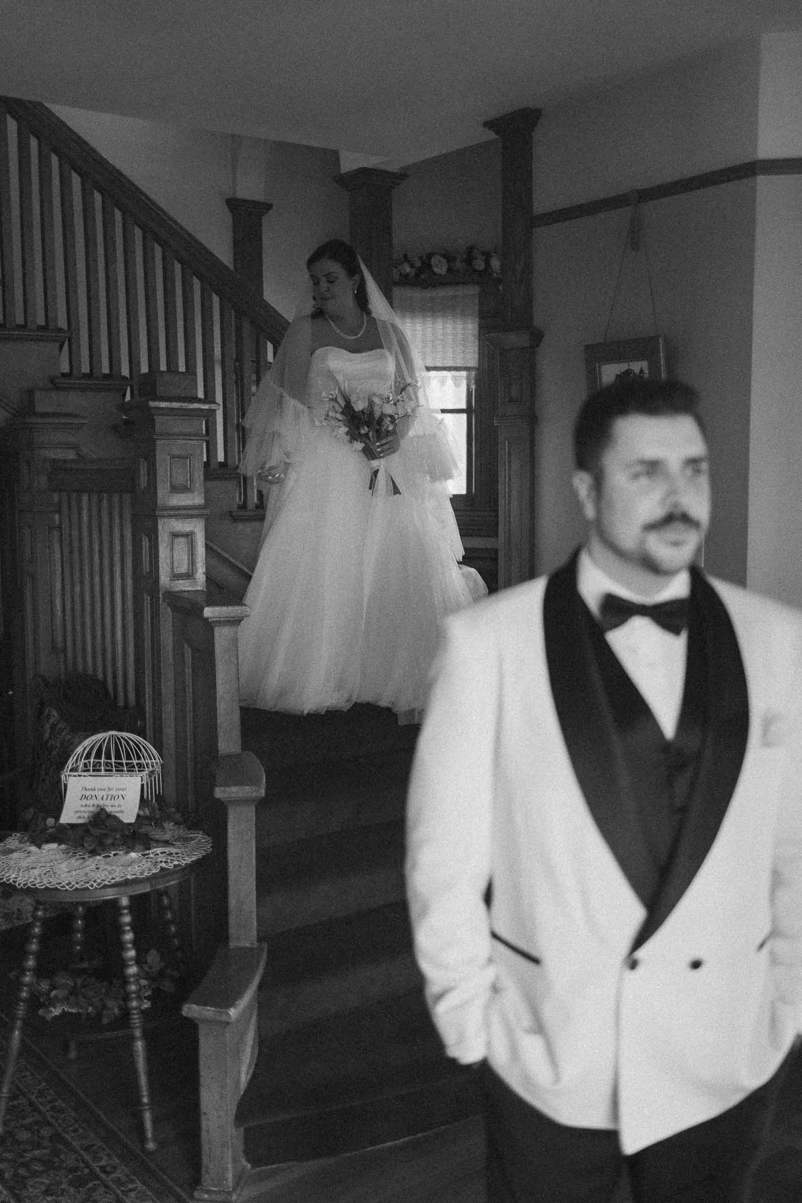  A bride in a wedding dress descends a staircase while holding a bouquet, as a groom in a light-colored tuxedo with a dark lapel stands in the foreground, slightly out of focus. 