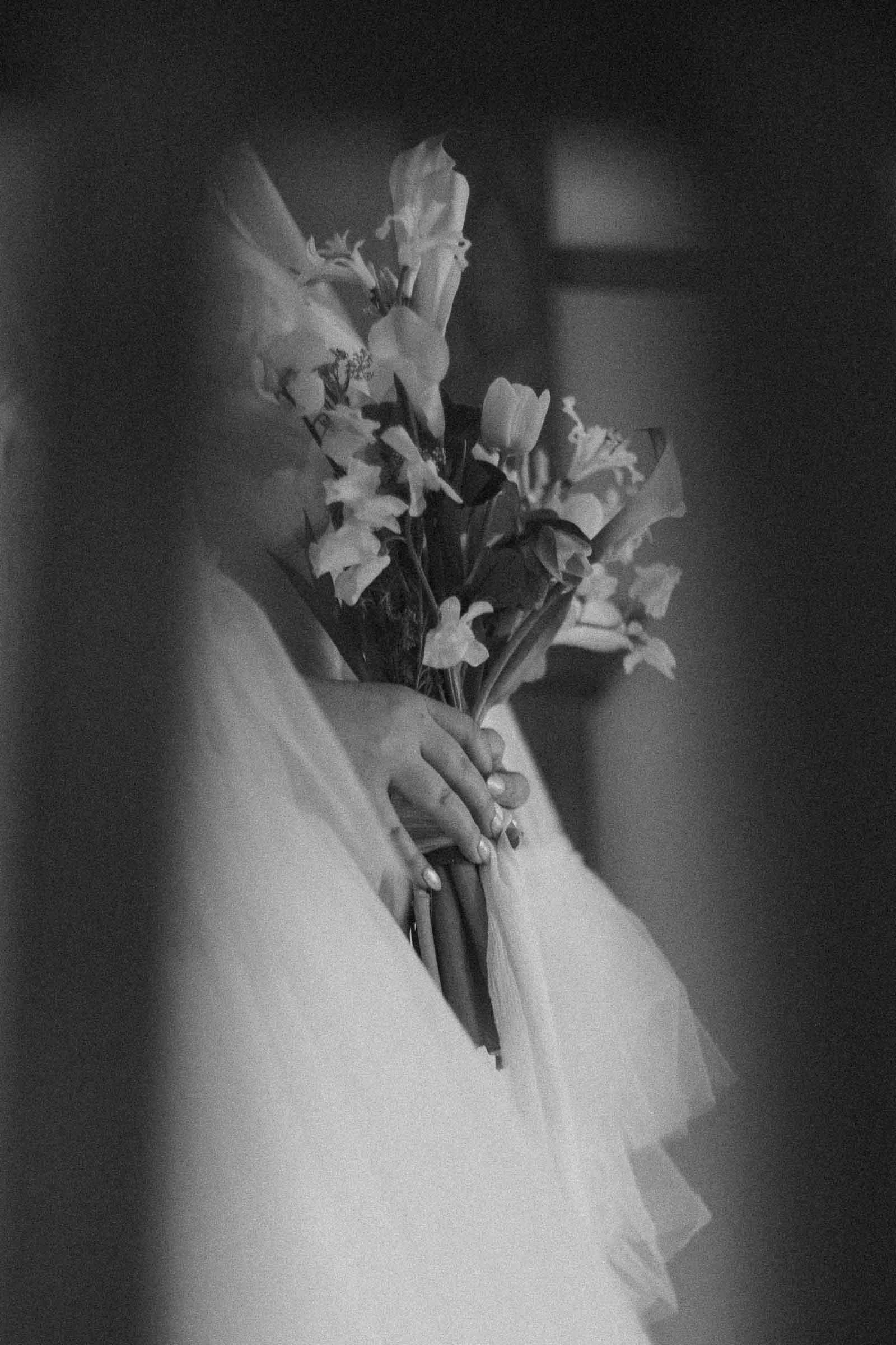  A black and white photo of a person holding a bouquet of flowers, with hands gently clasped around the stems. The person is partially obscured, wearing a light, flowing garment. The image is softly focused and framed by blurred foreground. 