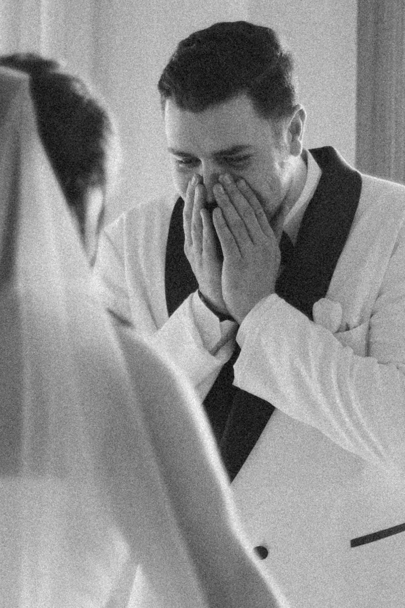  A groom in a white suit covers his mouth in joyful surprise, eyes crinkled with emotion, as he sees his bride, who is dressed in a veil and wedding gown, for the first time. 