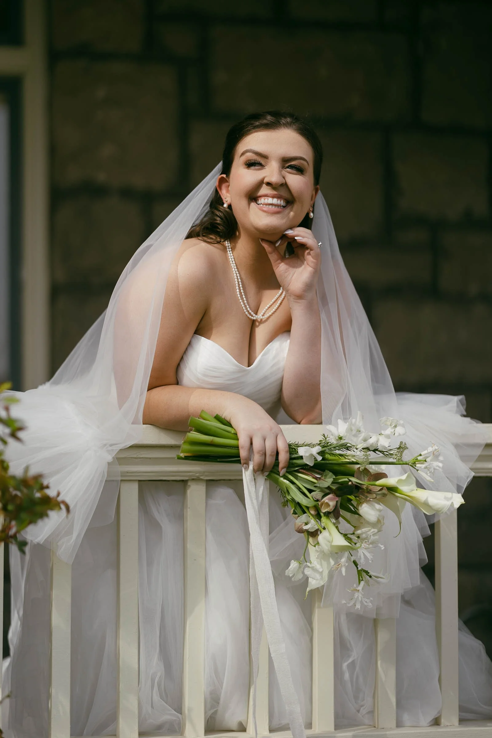  A smiling bride in a white dress and veil leans on a porch railing, holding a bouquet of white flowers and wearing a pearl necklace. A stone wall is visible in the background. 