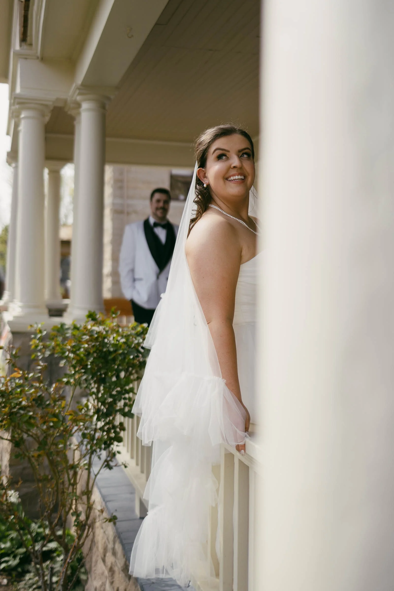  A bride in a white dress and veil stands on a porch, smiling and looking up. In the background, a groom in a white jacket and black bow tie stands, partially obscured by a column. 