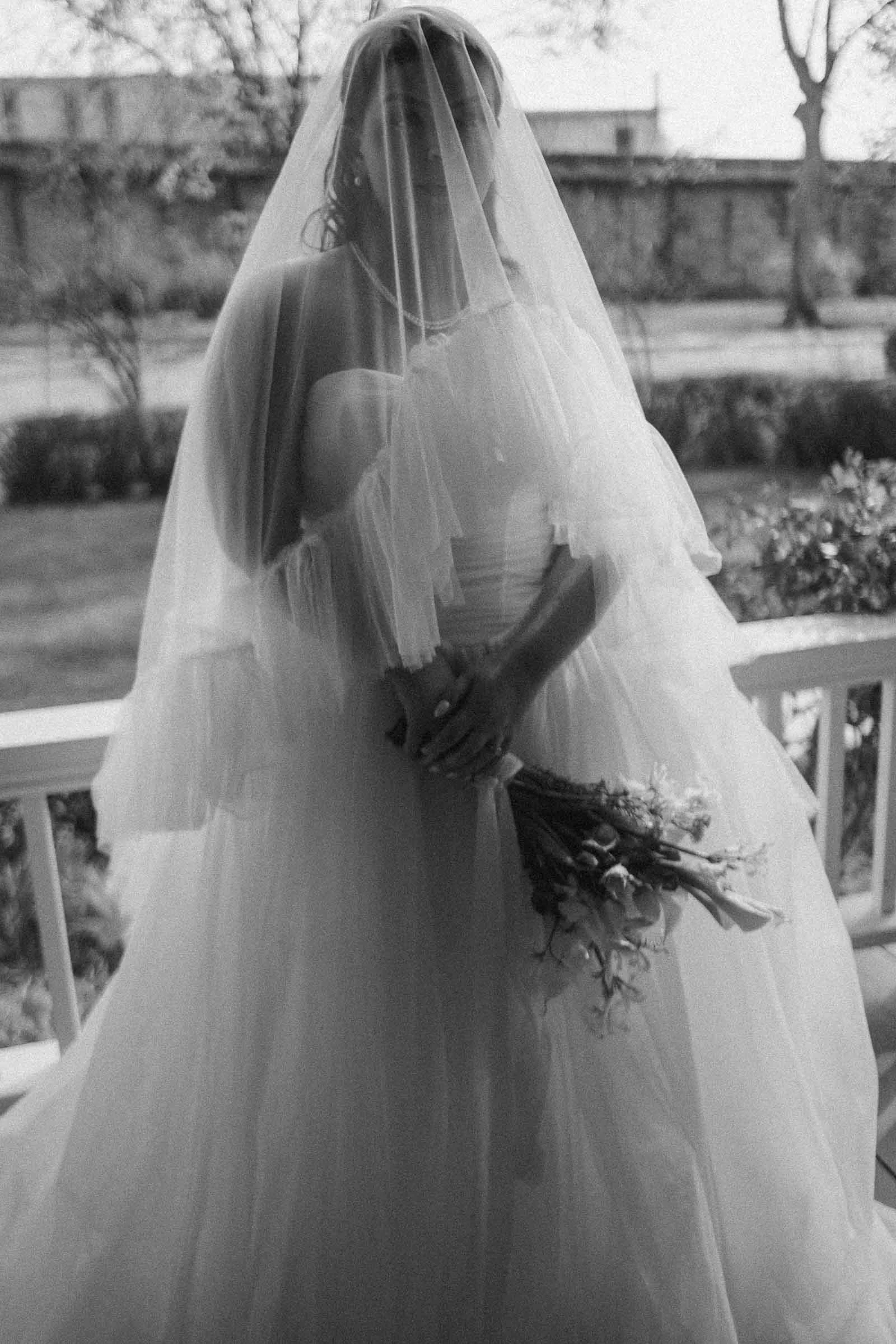  A bride in a strapless wedding dress and veil stands on a porch, holding a bouquet of flowers. The image is in black and white, giving it a timeless, classic feel. 