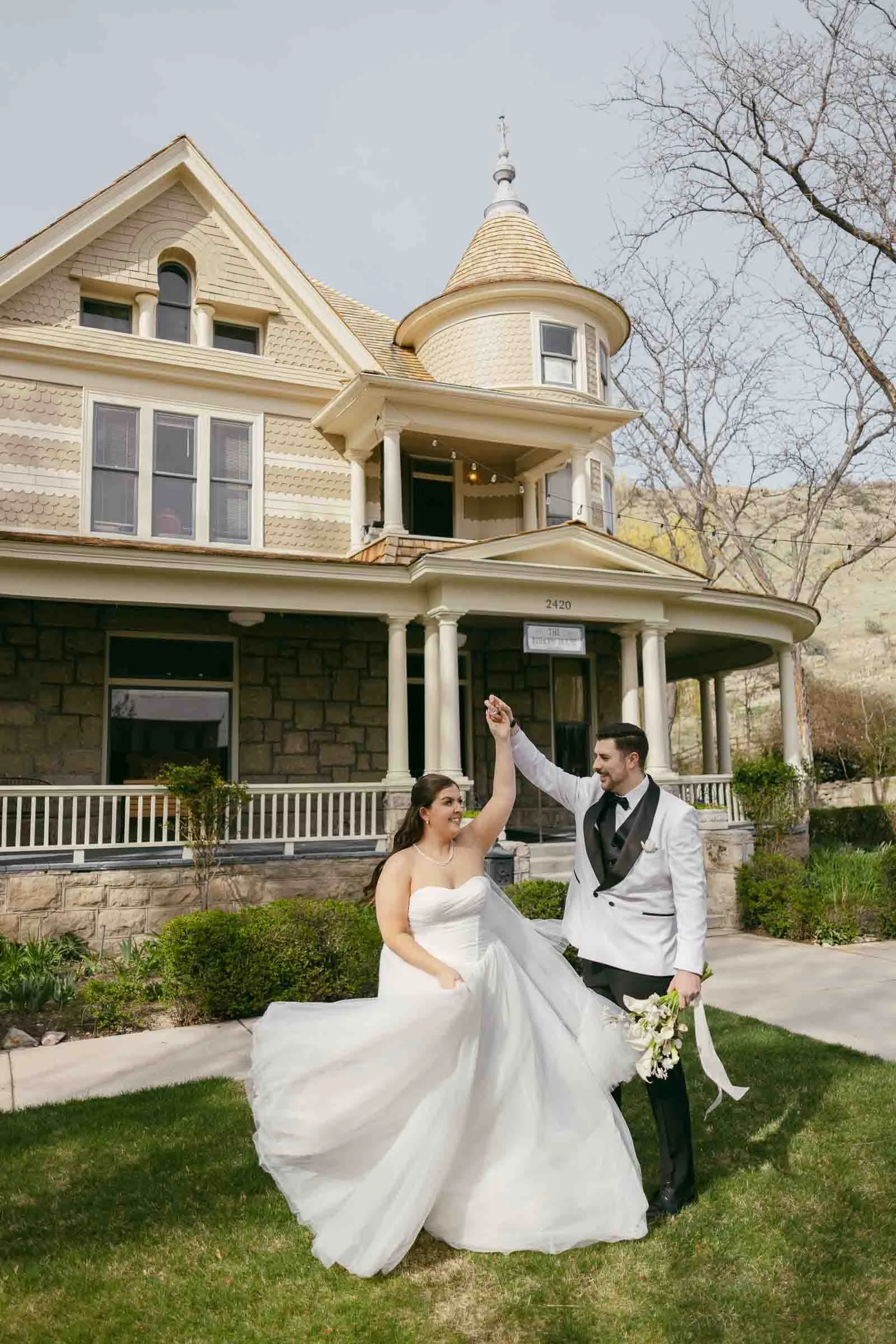  A bride and groom dance joyfully on a lawn in front of a large Victorian-style house. The bride's white dress fans out as she twirls, and the groom holds her hand and a bouquet, both smiling. 