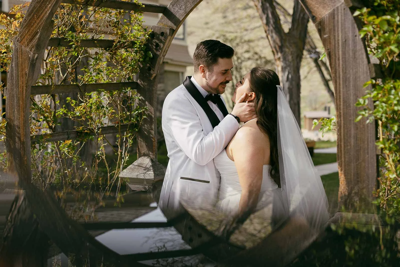 A groom in a white tuxedo gently holds his bride’s face as they smile at each other outside, framed by wooden garden arches and greenery. The bride wears a white dress and veil.