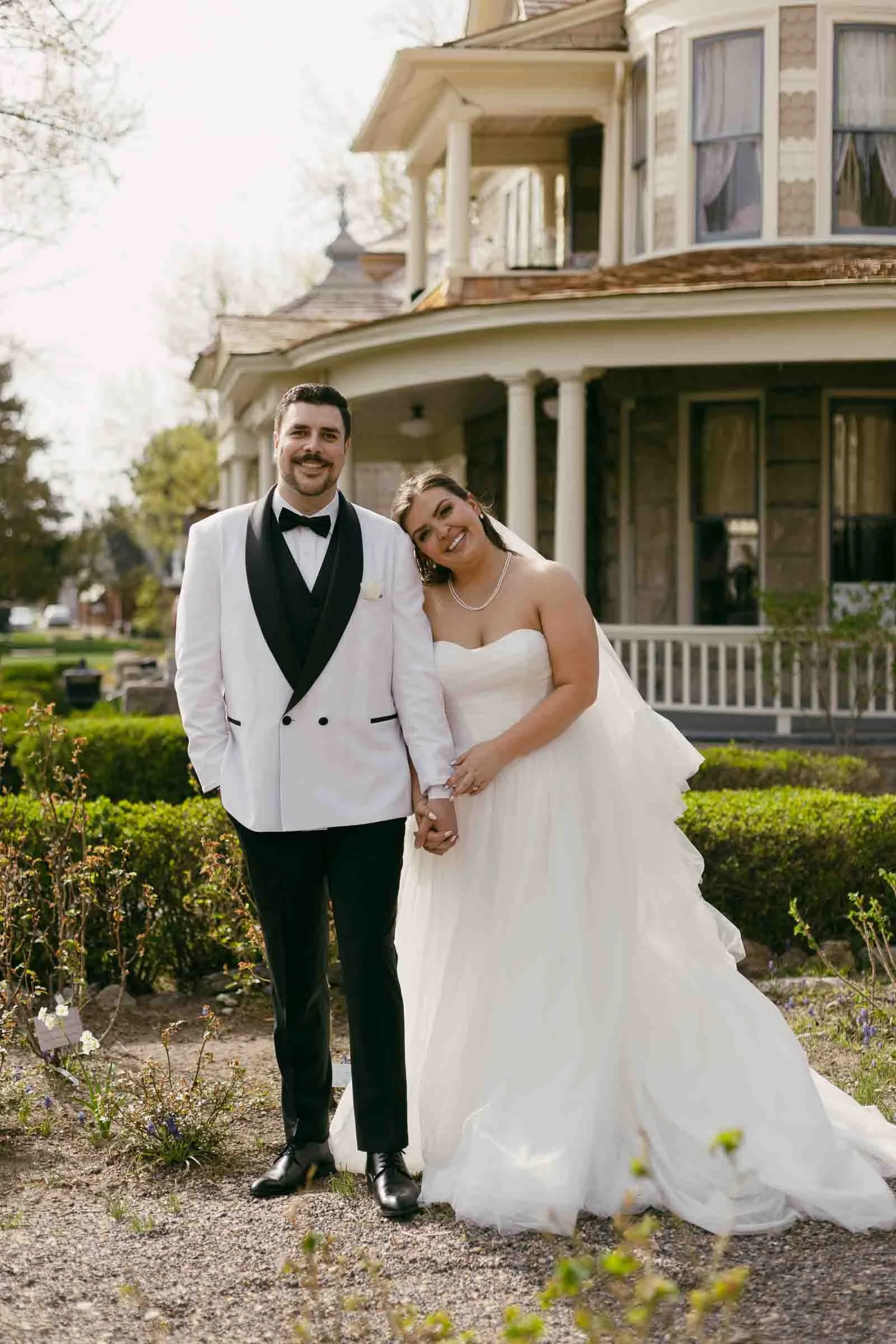 A bride in a white strapless gown leans her head on the groom’s shoulder. The groom wears a white tuxedo jacket with black lapels. They stand outdoors, holding hands, in front of The Bishops House.
