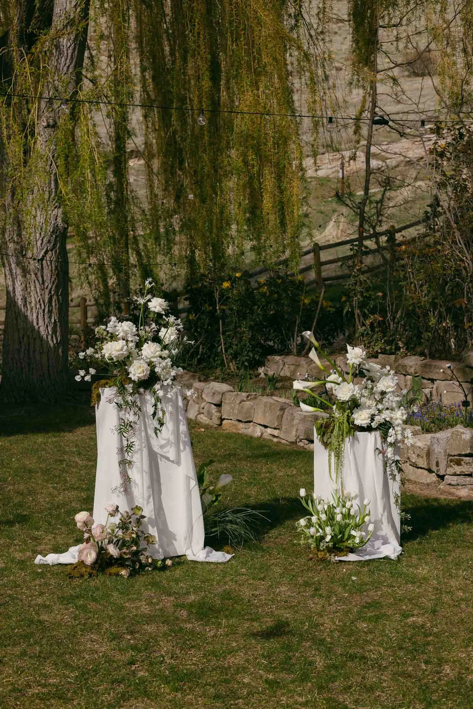  Two white-draped pedestals topped with lush white floral arrangements stand on grass; additional flowers and greenery surround their bases, with a stone wall, trees, and string lights in the background. 
