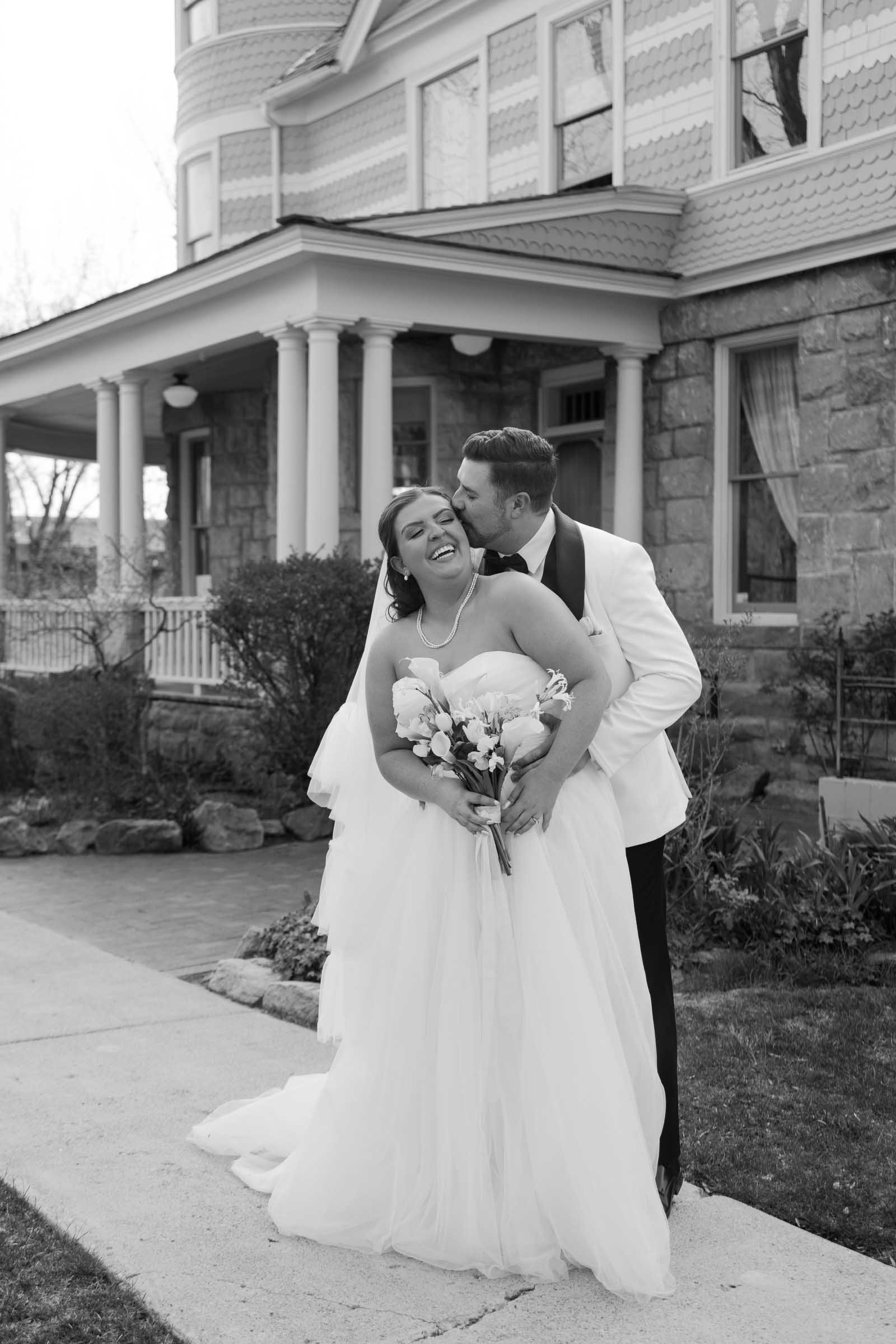  A bride and groom stand smiling in front of a large house. The groom, in a white jacket, embraces the bride from behind and kisses her cheek. The bride holds a bouquet and wears a strapless gown. The photo is in black and white. 
