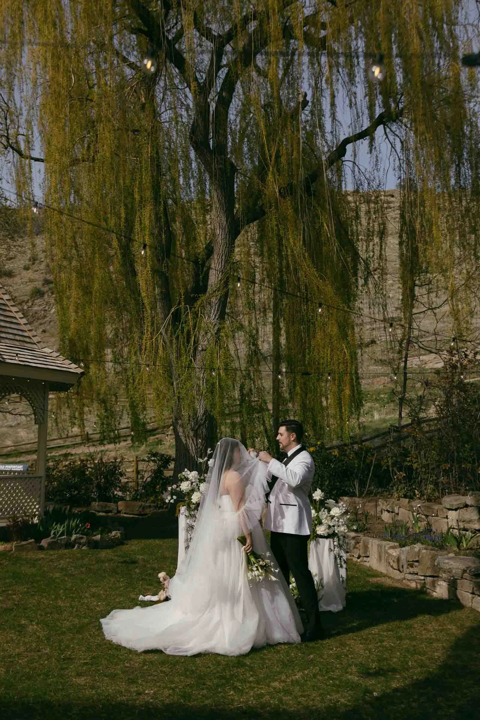  A bride and groom stand together outdoors under a large willow tree during their wedding ceremony. The groom adjusts the bride’s veil, and they are surrounded by greenery and floral arrangements. 