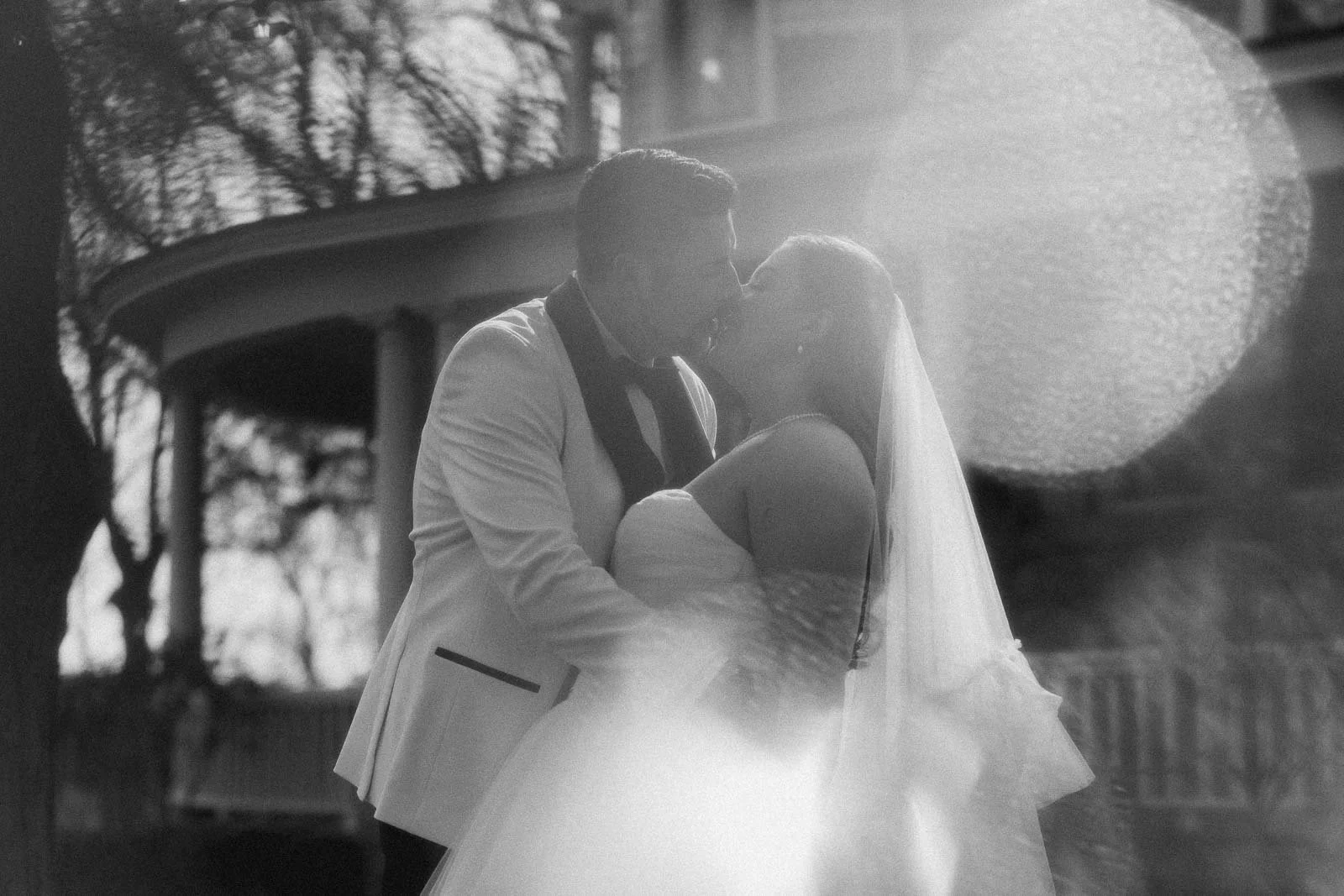 A bride and groom share a kiss outdoors in front of The Bishops House. The wraparound porch is in the background. Sunlight and lens flare create a dreamy, romantic effect in the black-and-white image.