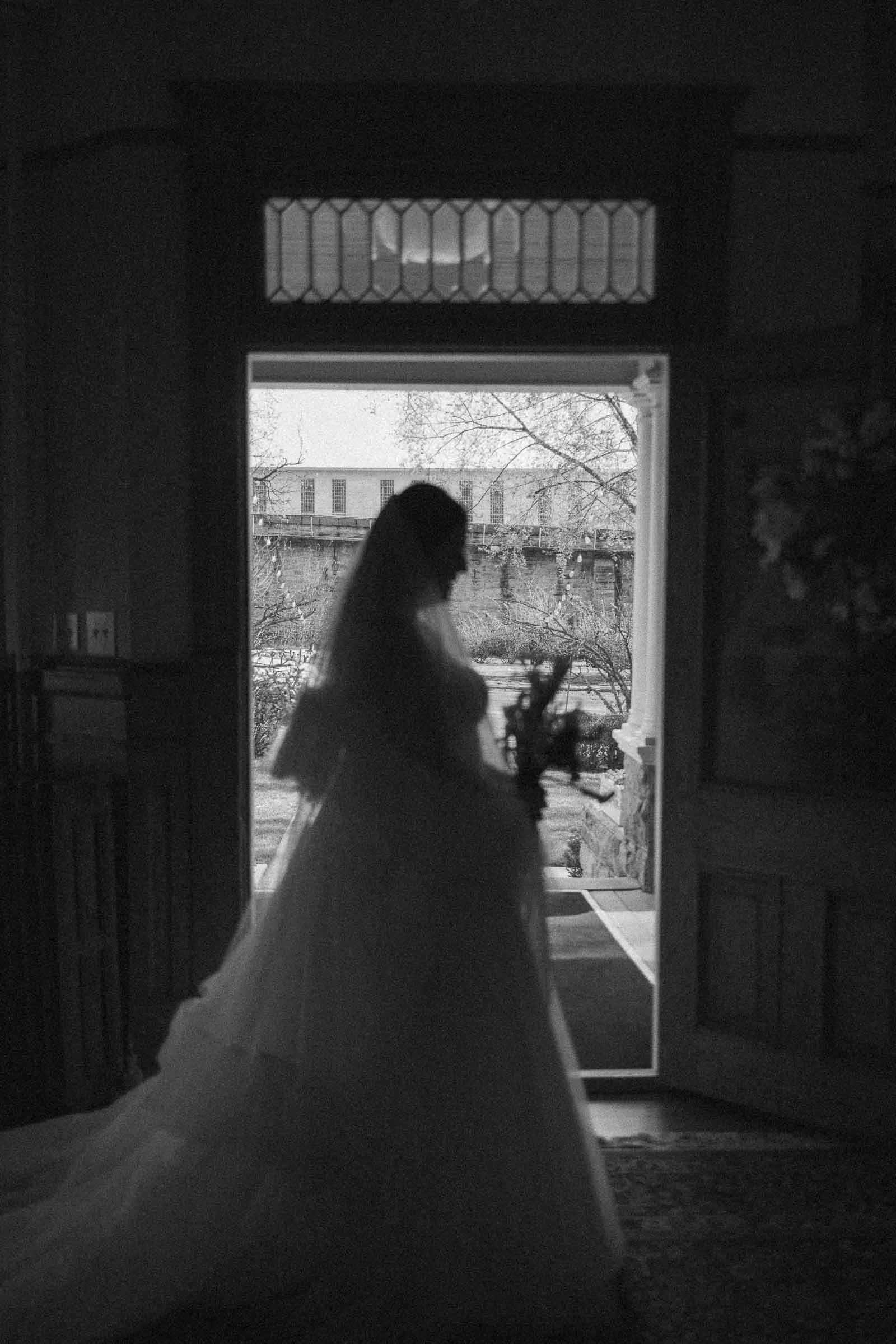  A silhouetted bride holding a bouquet stands inside by an open door, with soft daylight illuminating her figure against an outdoor backdrop. 