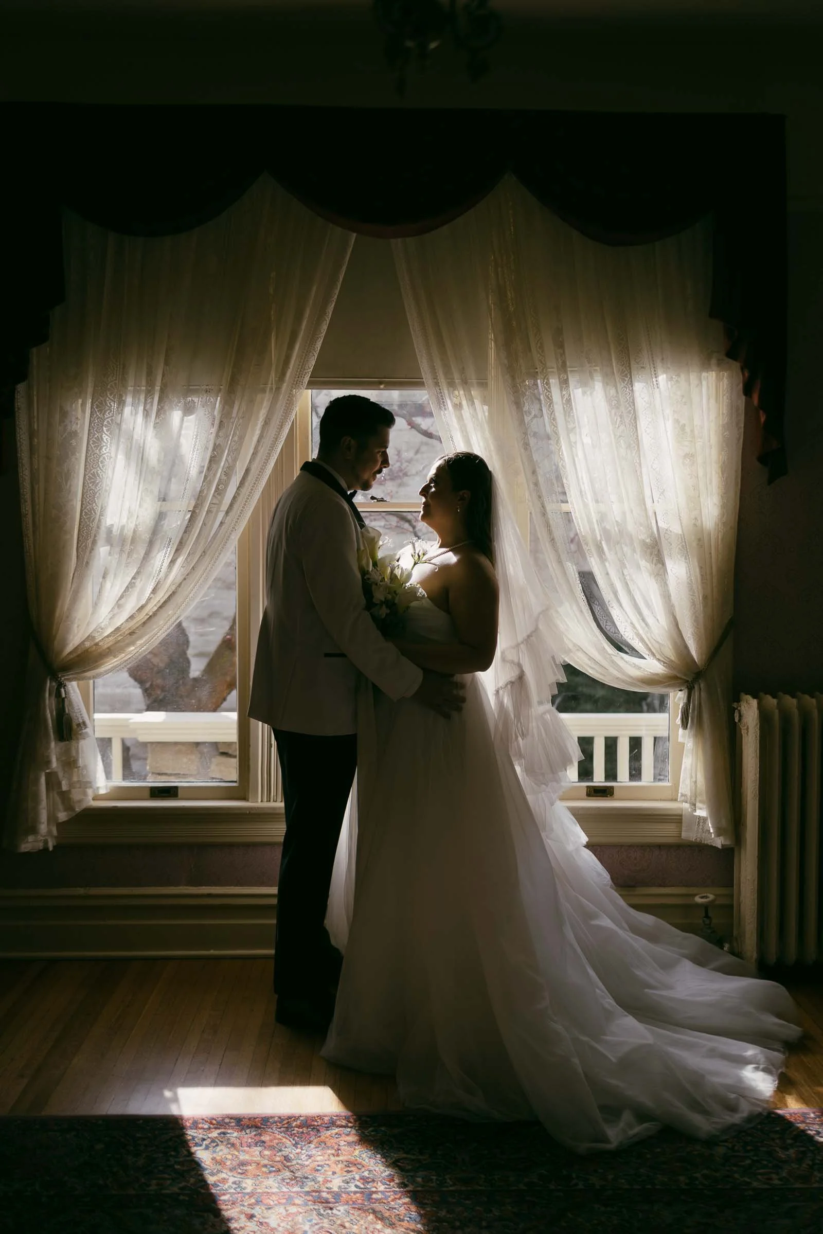  A bride and groom stand facing each other in front of a sunlit window with sheer curtains, embracing and holding a bouquet. They are silhouetted by the natural light coming through the window. 