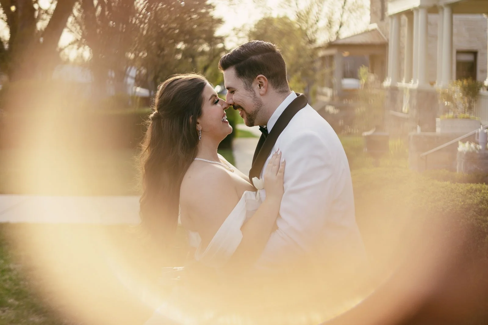 A bride and groom smile and lean in close together outdoors, framed by a soft, golden light effect. The bride wears an off-shoulder white dress, and the groom wears a white tuxedo jacket with black details.