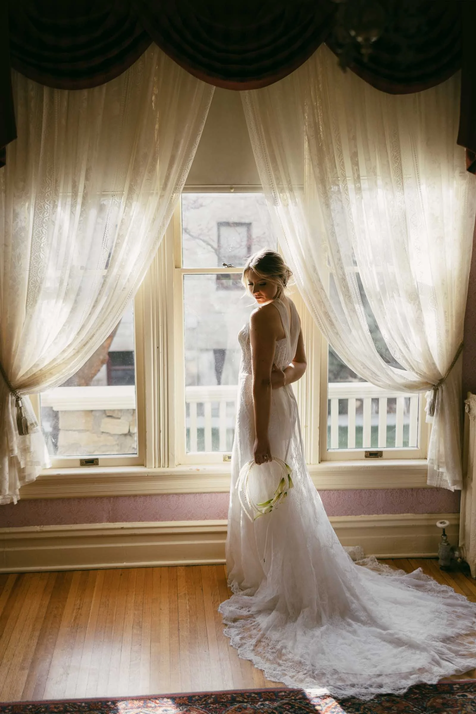  A bride in a white lace gown stands by a large window with sheer curtains, holding a small bouquet and looking over her shoulder. Sunlight streams in, illuminating her and the wooden floor. 