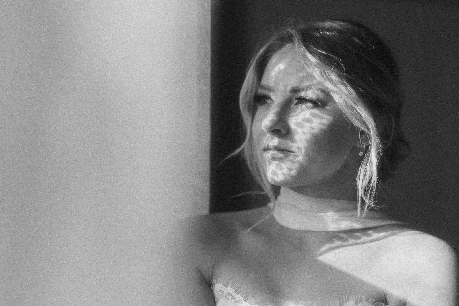  Black and white photo of a woman with light patterns on her face, looking thoughtfully into the distance. She has light hair pulled back and wears a strapless top with a soft expression. 