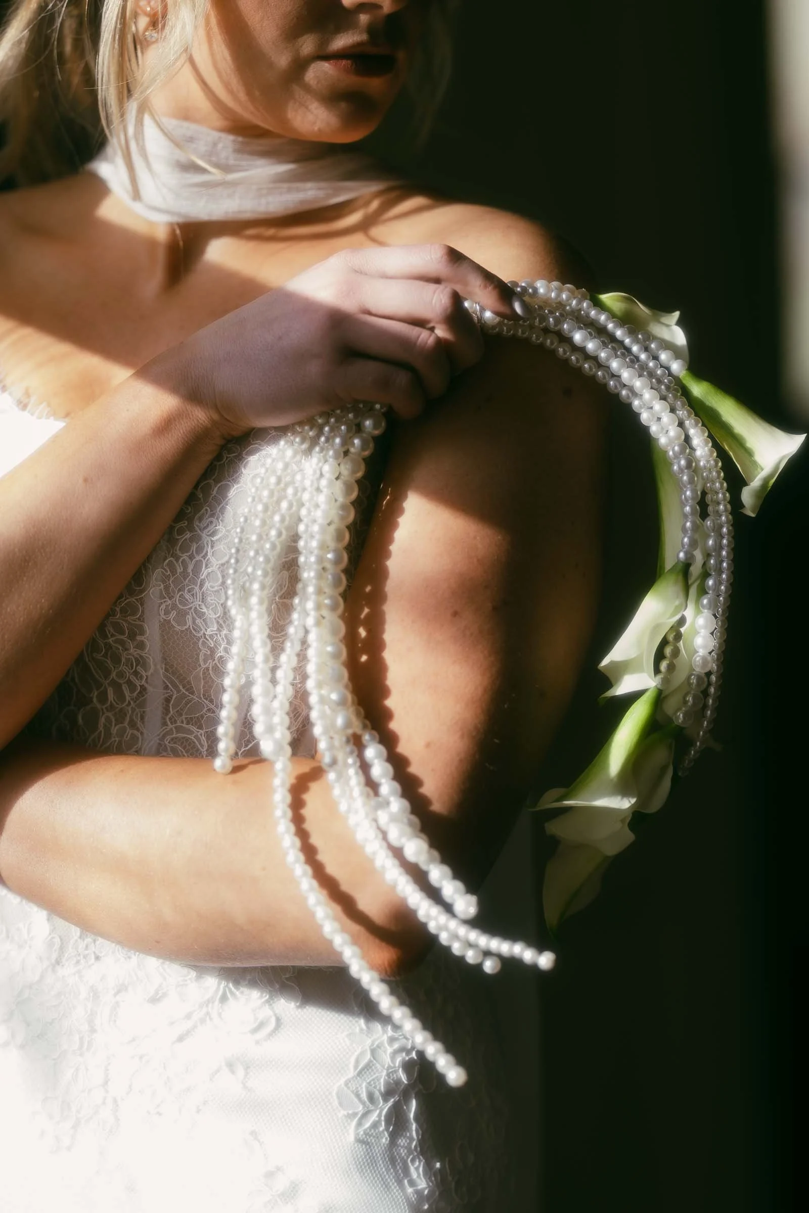  A woman in a white lace dress holds calla lilies and strands of pearls draped over her arm, with soft sunlight highlighting her features and accessories. 