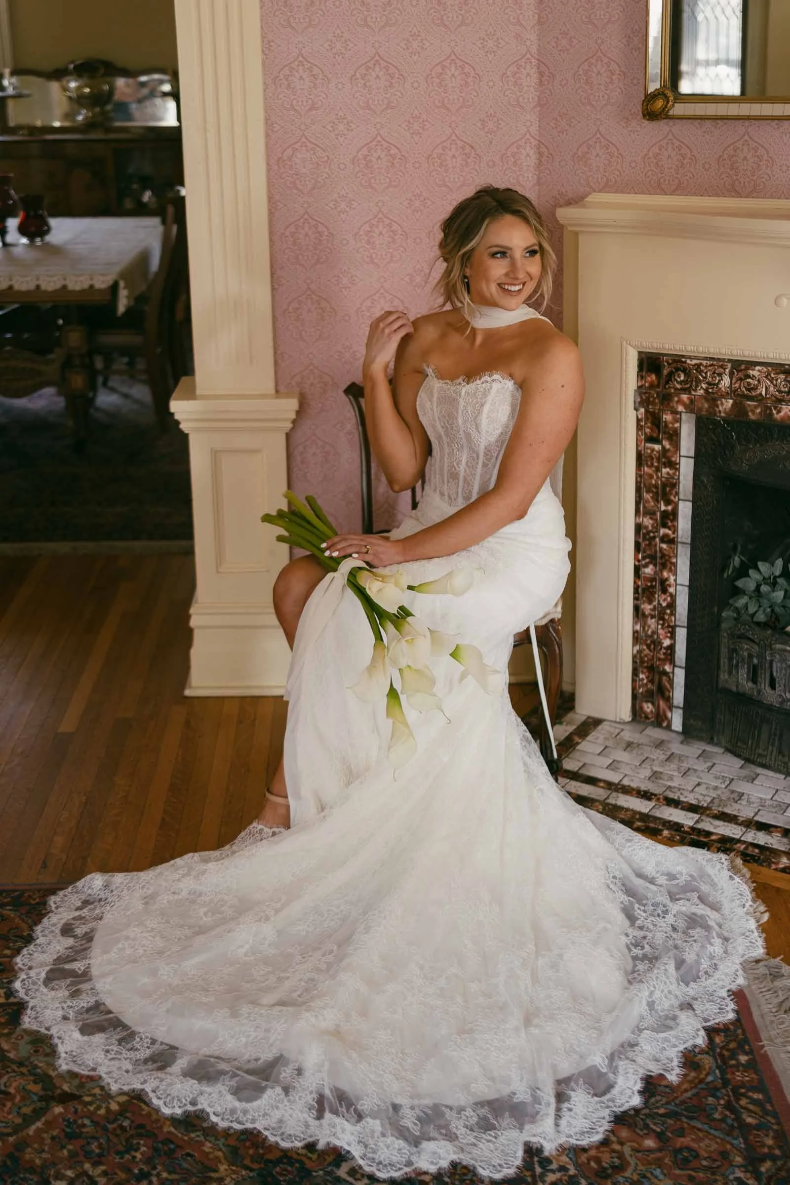  A bride in a strapless white lace wedding gown sits on a chair near a fireplace, holding a bouquet of white calla lilies and smiling. The setting is a warmly lit, vintage-style room with wooden floors and pink wallpaper. 