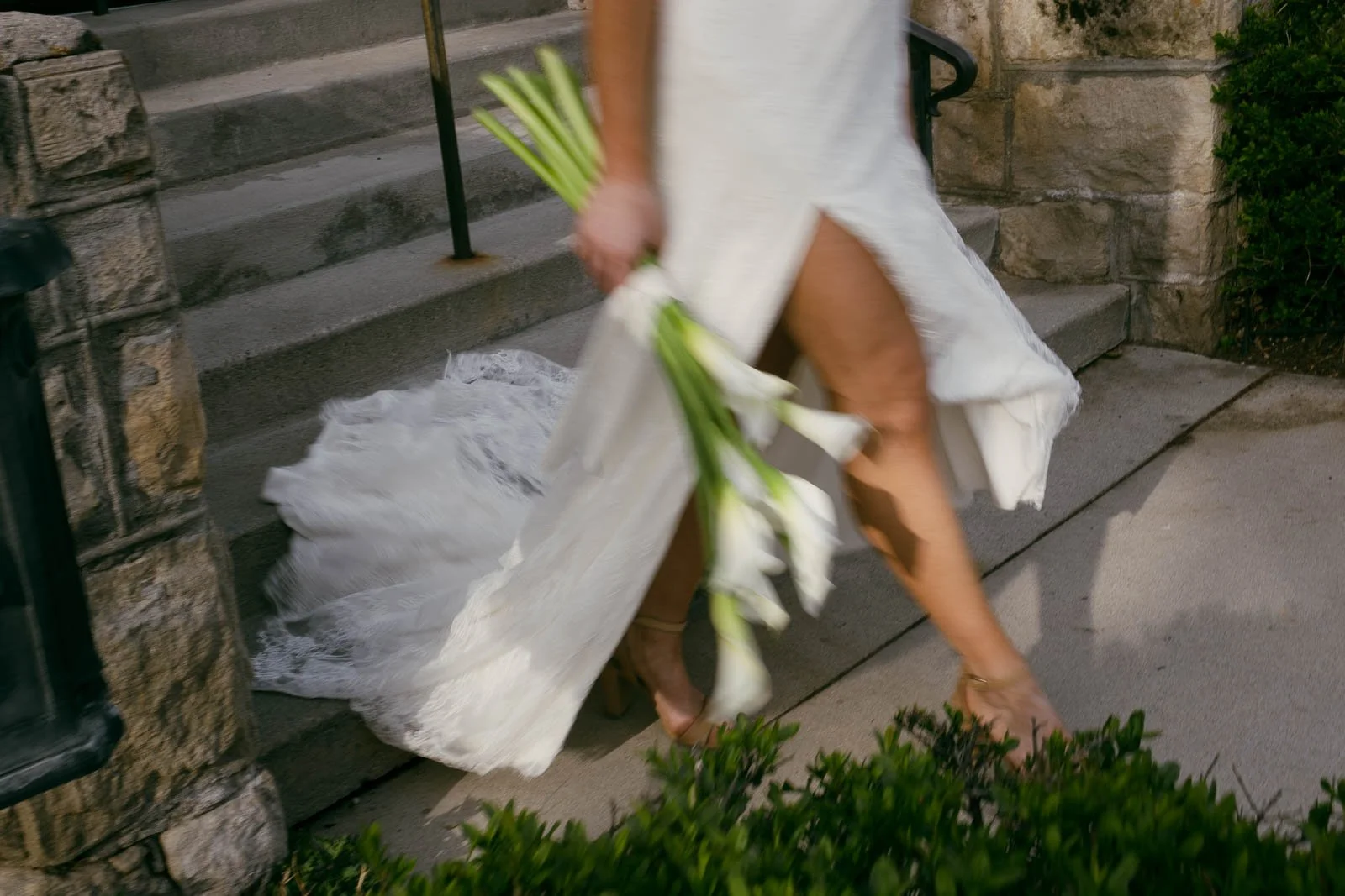  A person in a flowing white dress walks down stone steps, holding a bouquet of white calla lilies. Their face is out of frame, and greenery borders the walkway. 