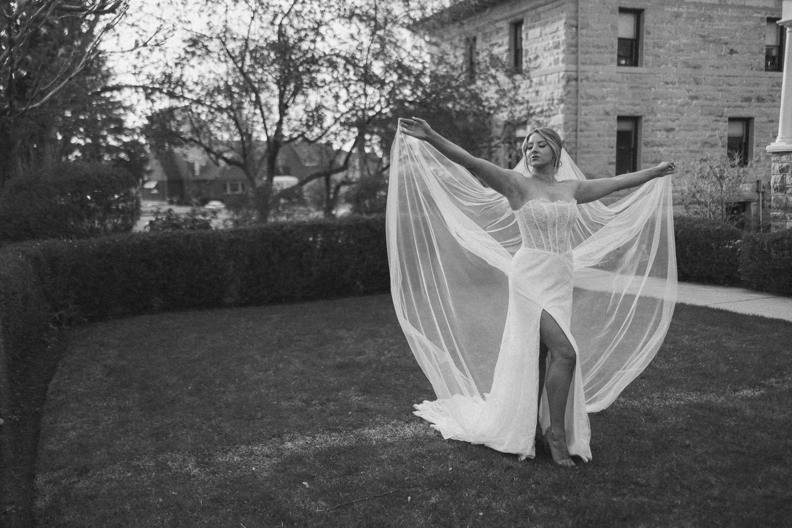  A woman in a strapless wedding dress stands on grass, holding her veil out wide with both arms. She poses gracefully in front of a stone building and trees. The image is in black and white. 