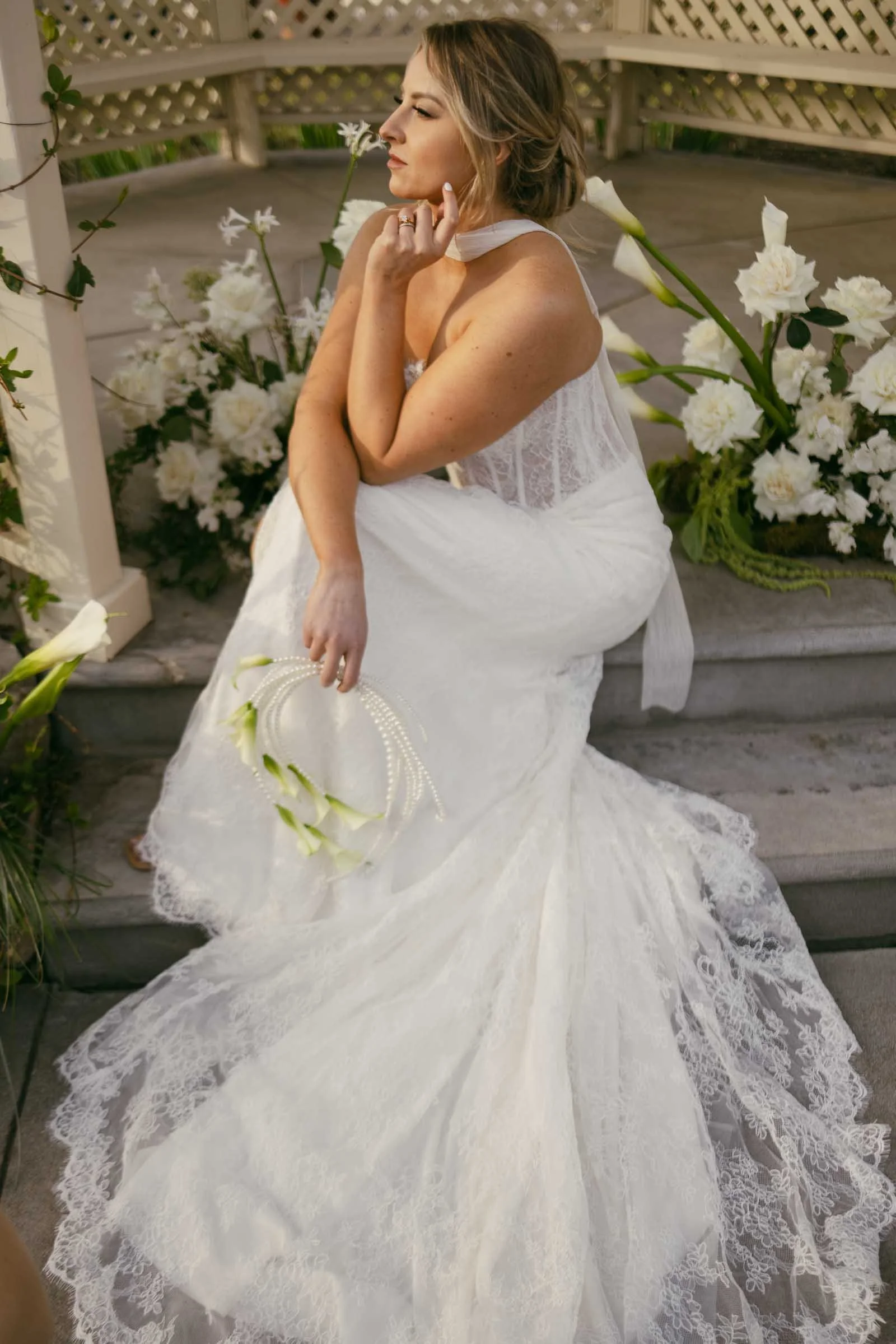  A woman in a lace wedding dress sits on outdoor steps beside white flowers, resting her chin on her hand and gazing thoughtfully into the distance while holding a small bouquet. 
