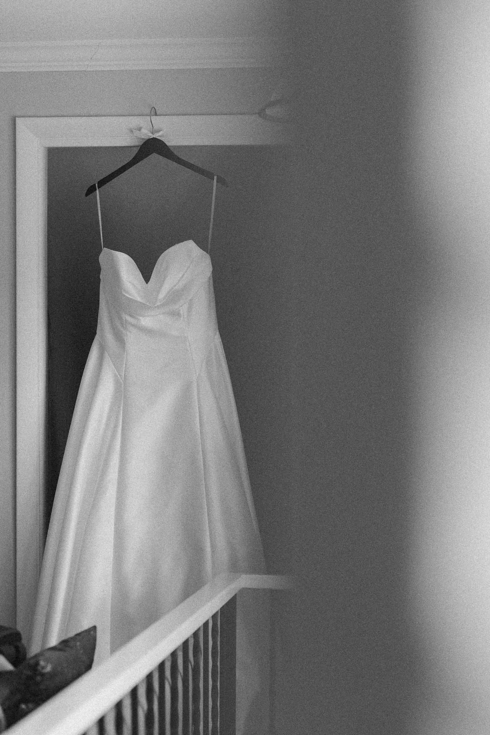  A strapless white wedding dress hangs on a hanger in a doorway, with soft lighting and a blurred foreground partially obscuring the view. 