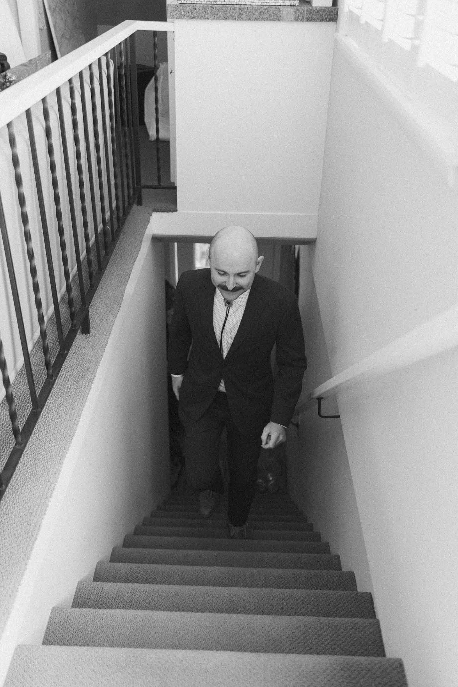  A man in a suit with a mustache walks up a carpeted staircase inside a house, looking down. The photo is in black and white. 