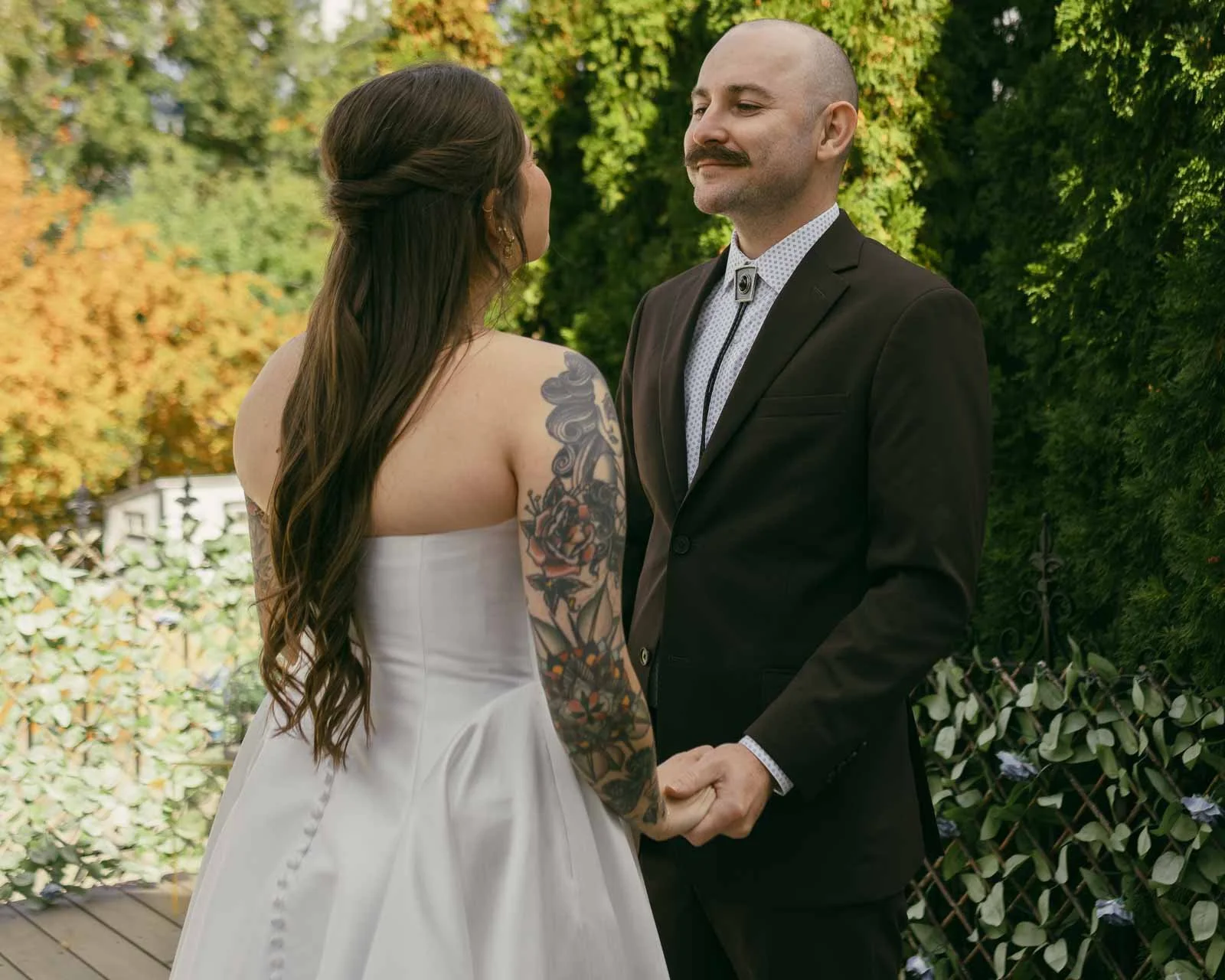  A bride in a white dress with long hair and a tattooed arm holds hands with a groom in a dark suit and bolo tie. They stand facing each other outdoors, surrounded by greenery and autumn trees. They have just had a first look and are celebrating thei