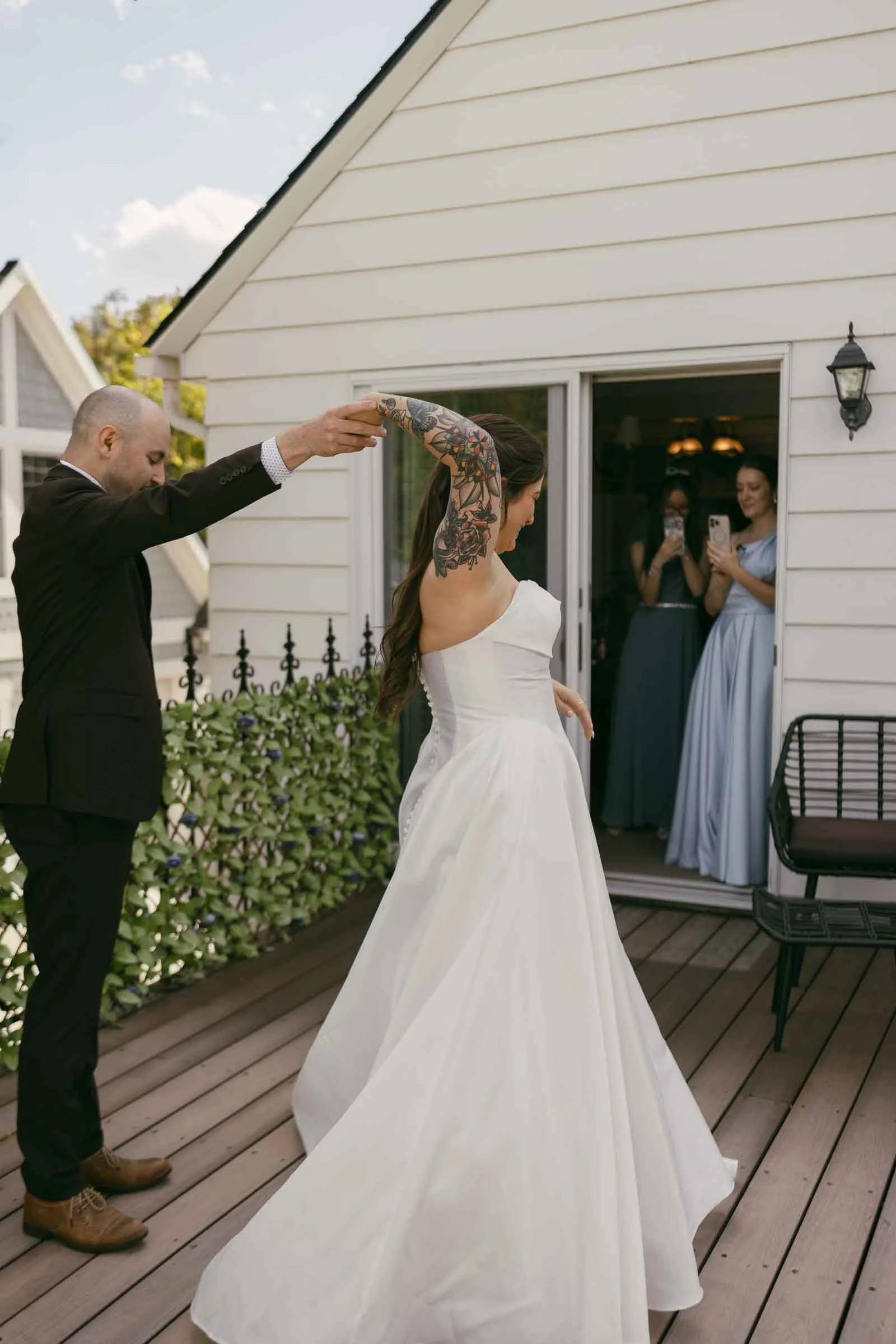  A bride in a white dress twirls with a man in a suit on a wooden deck, while two women, the bride’s sister and niece, in blue dresses watch and take photos from inside the house. 