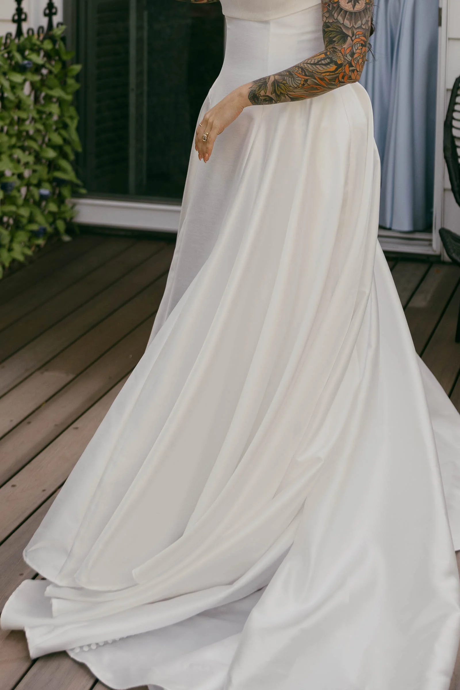  A bride in a sleeveless white wedding dress stands on a wooden deck, showing detailed tattoos on their arm. The dress features a full, flowing skirt with elegant draping. Greenery and a blue dress are visible in the background. 