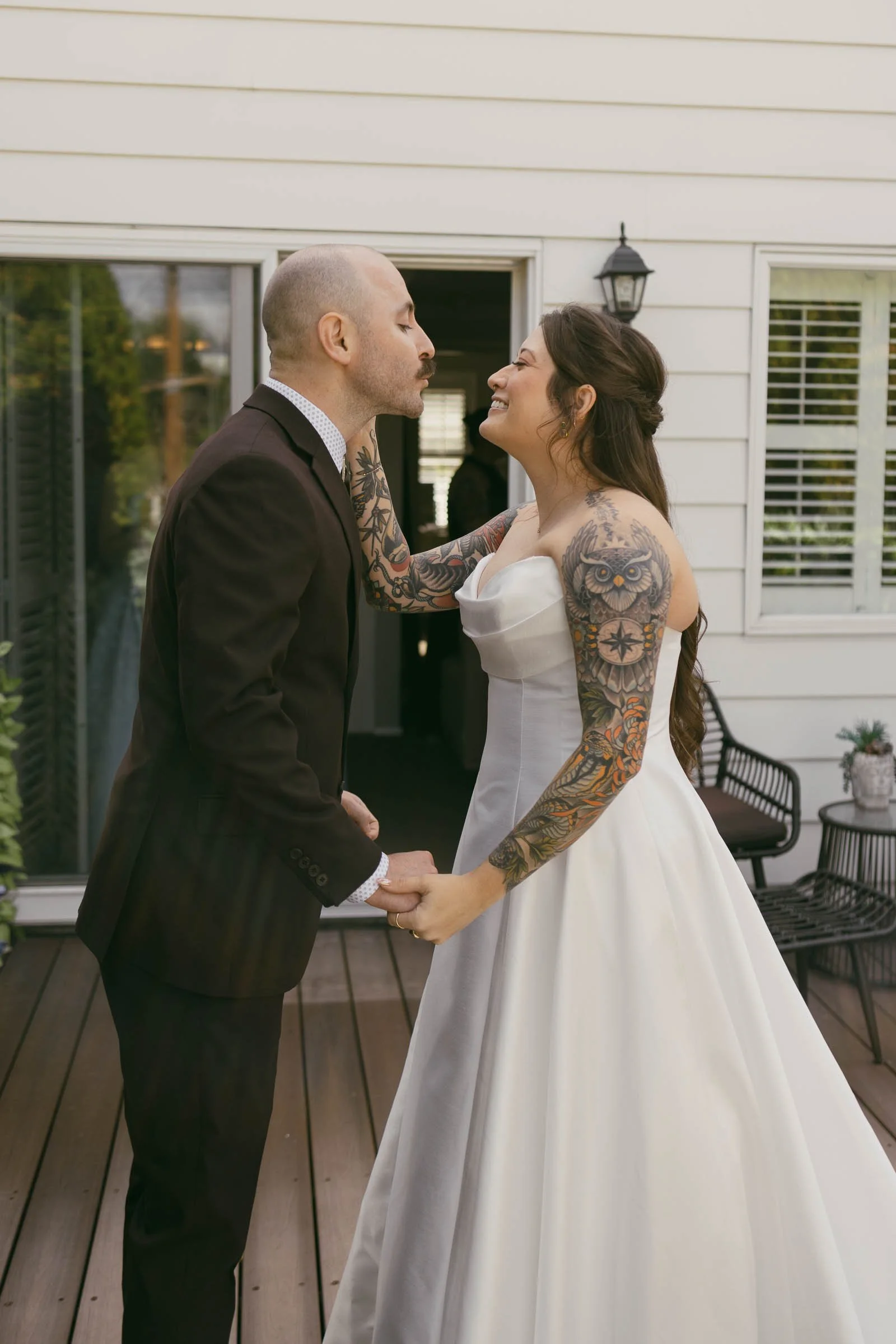  A bride in a white dress with colorful arm tattoos and a groom in a brown suit smile at each other on a wooden deck, holding hands. The bride gently touches the groom’s face outside a white house. 