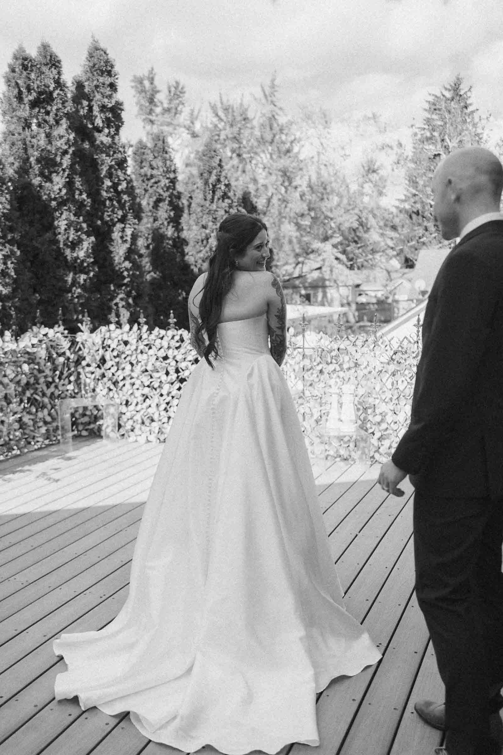  A bride in a long white gown smiles over her shoulder while standing on a wooden deck outdoors. A man in a suit stands nearby. Trees and foliage are visible in the background. The photo is in black and white. 