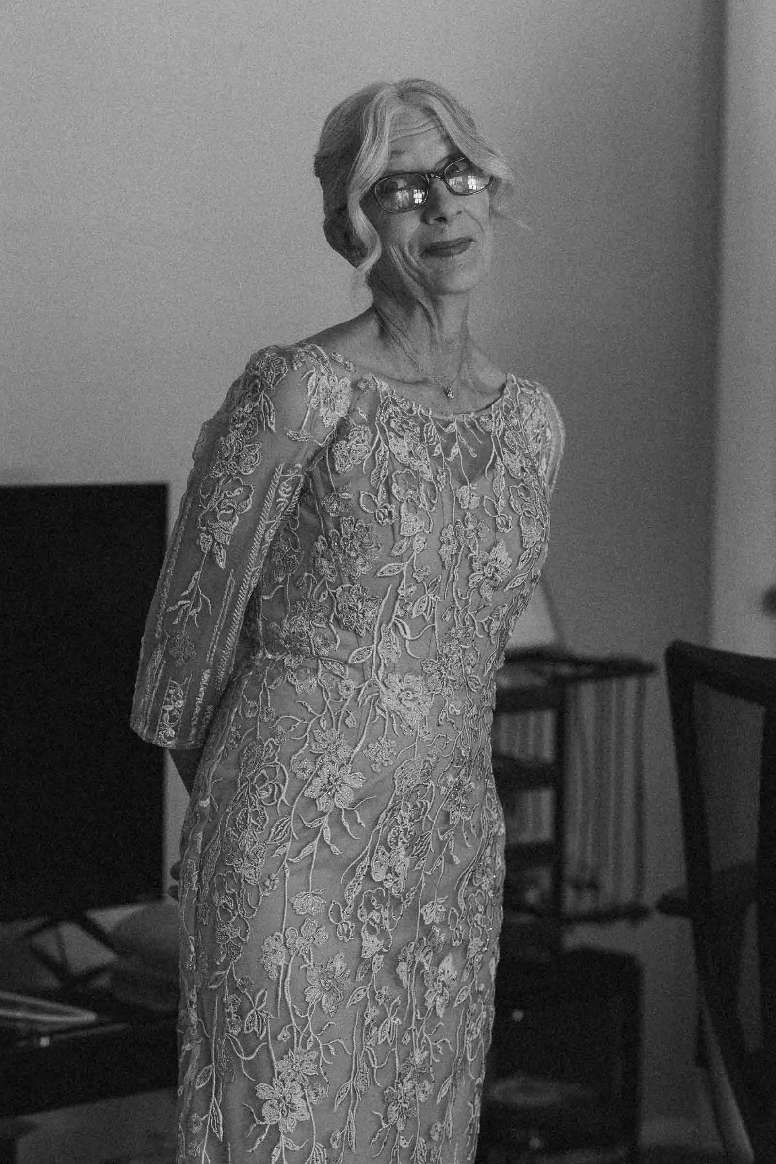 The bride’s mother, with glasses and light hair, styled in an updo, wears an ornate, embroidered lace dress. She stands indoors, smiling softly, with her hands behind her back. The photo is in black and white. 