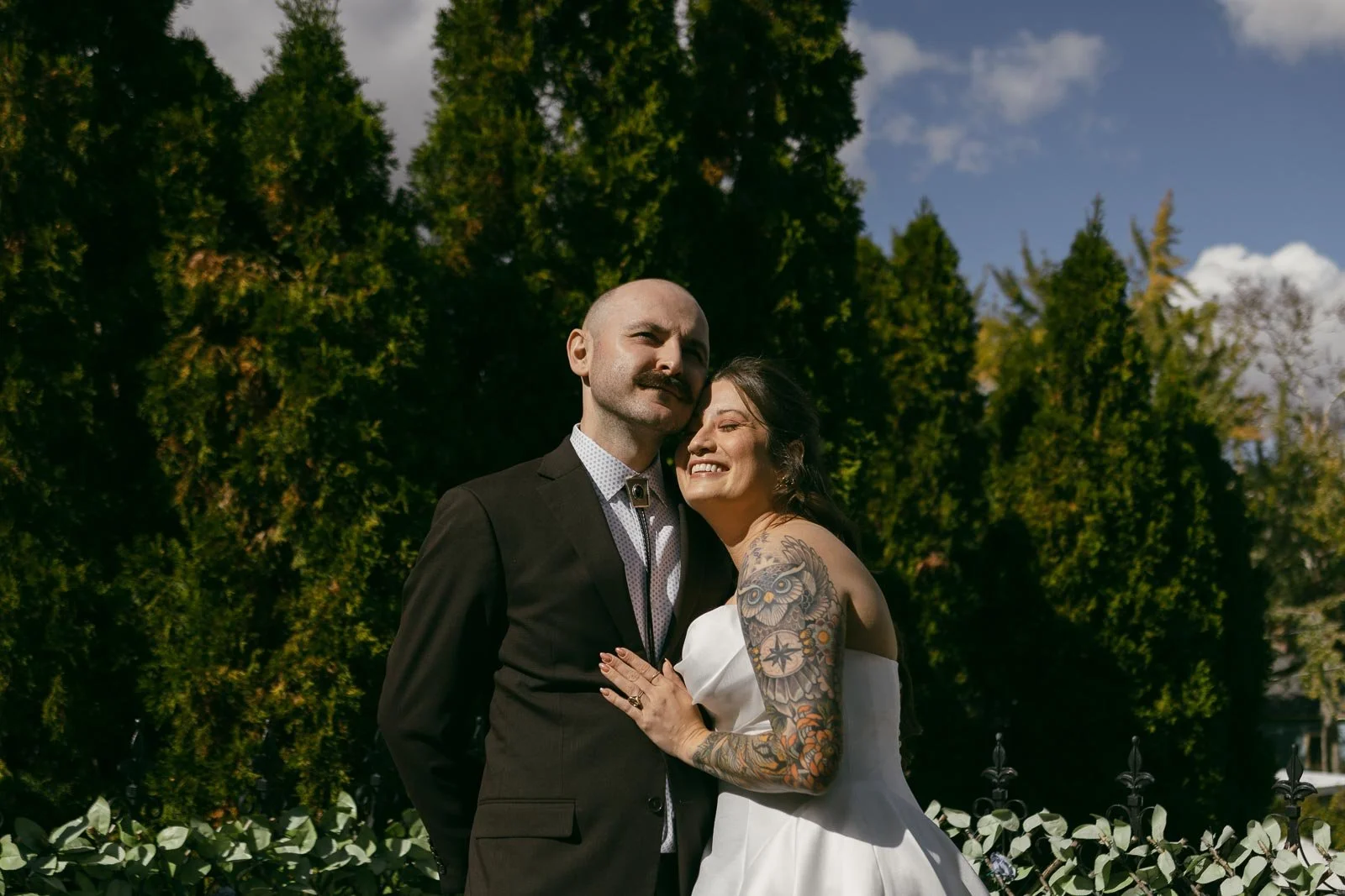  A bride in a white dress with visible tattoos smiles, embracing a groom in a dark suit and tie. They stand outdoors in front of tall green trees under a partly cloudy sky. 