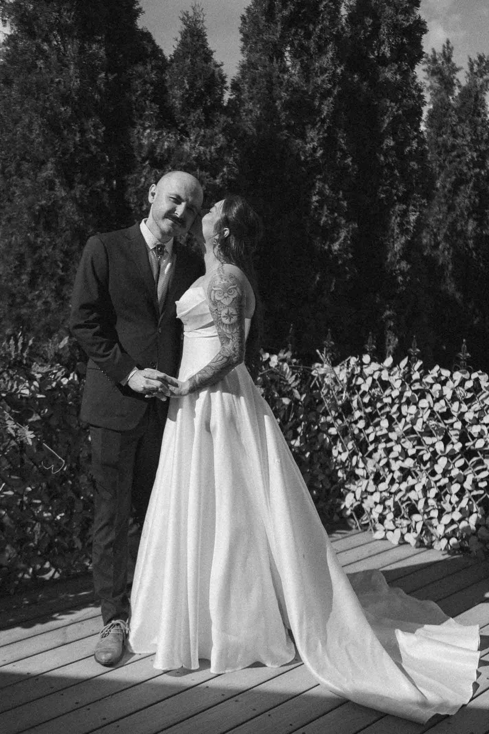  A joyful couple stands outdoors, holding hands. The groom wears a dark suit, and the bride, with visible tattoos, wears a long white dress and kisses his cheek. Tall trees and foliage are in the background. 