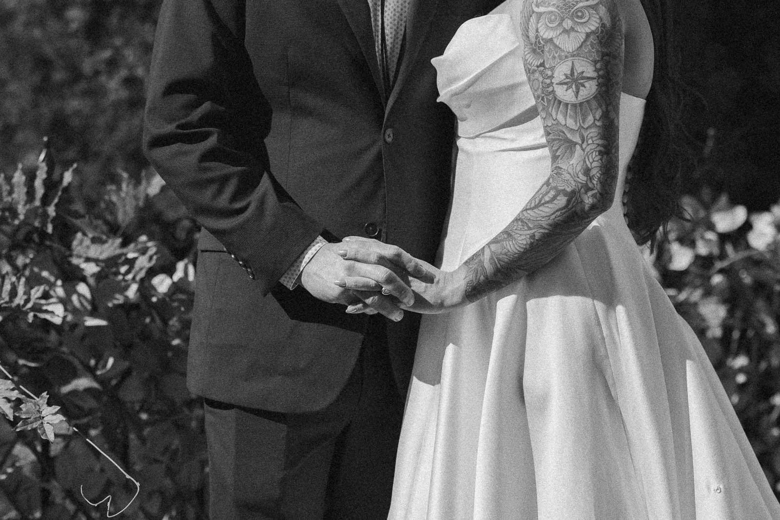  A black and white photo of a couple holding hands outdoors; the person in a suit stands next to another in a strapless dress with a tattooed arm. Their faces are out of frame, and foliage is in the background. 