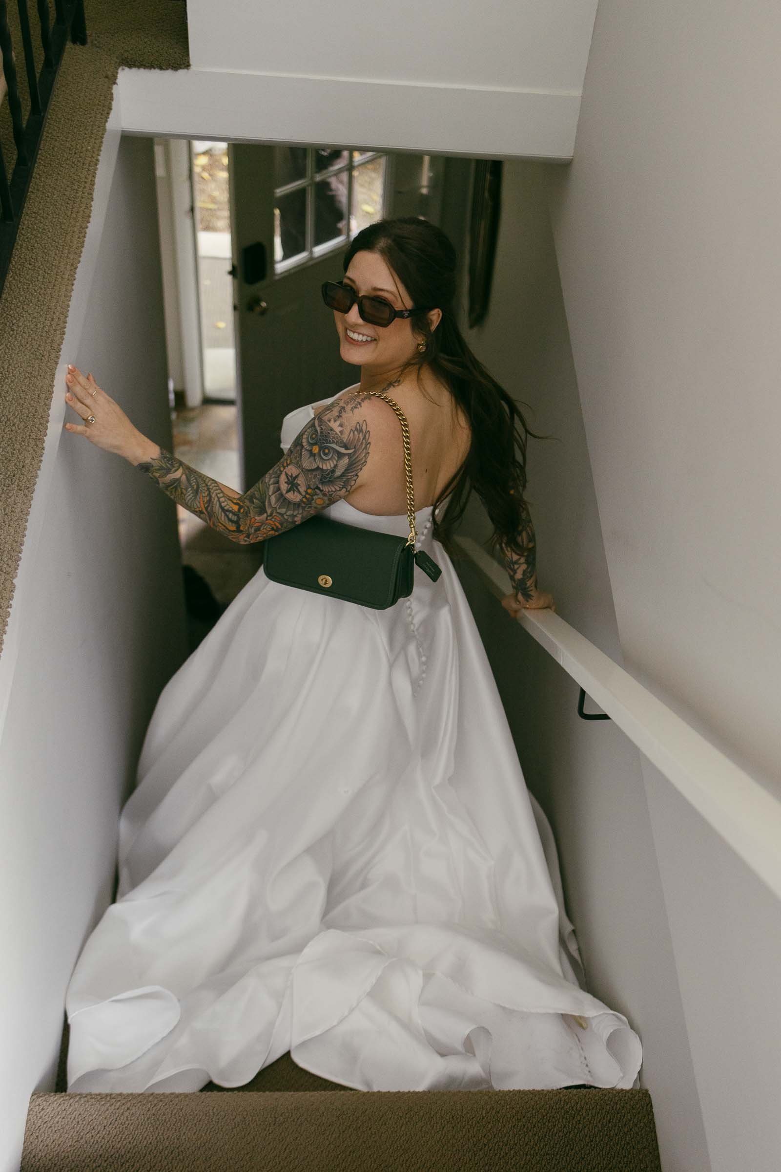  A smiling bride with long hair, sunglasses, tattoos, and a white wedding dress walks down a staircase, holding the handrail and a black purse over her shoulder. 