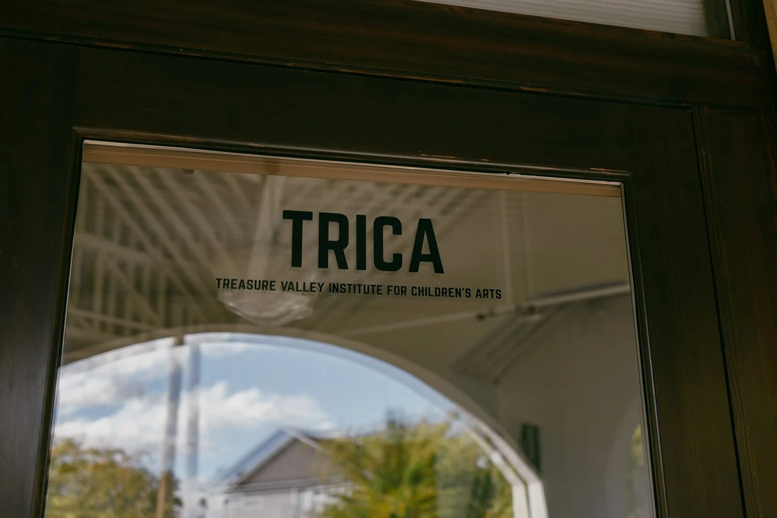 Glass door with the text "TRICA" and "Treasure Valley Institute for Children's Arts" in black lettering, reflecting an outdoor scene with trees, a house, and a partly cloudy sky. 