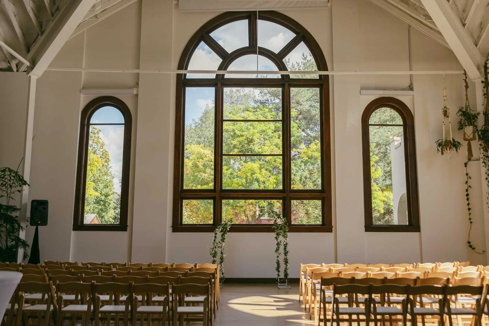  Sunlit wedding ceremony space with rows of wooden chairs facing a large arched window. Greenery decorates a simple arch, and trees are visible outside through the windows. The room has high, white-beamed ceilings. 