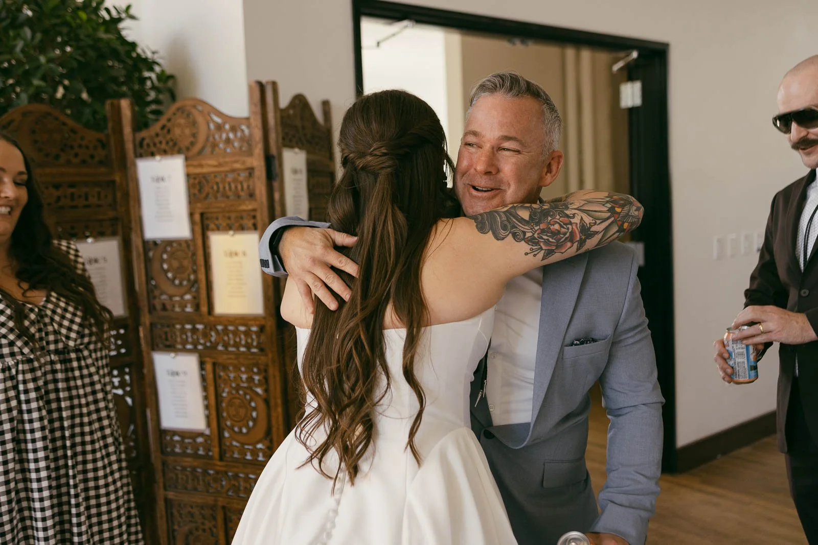  A bride in a white dress hugs a smiling man in a light blue suit at an indoor event. Other guests stand nearby, one holding a drink, in a warmly lit room with decorative wooden panels. 