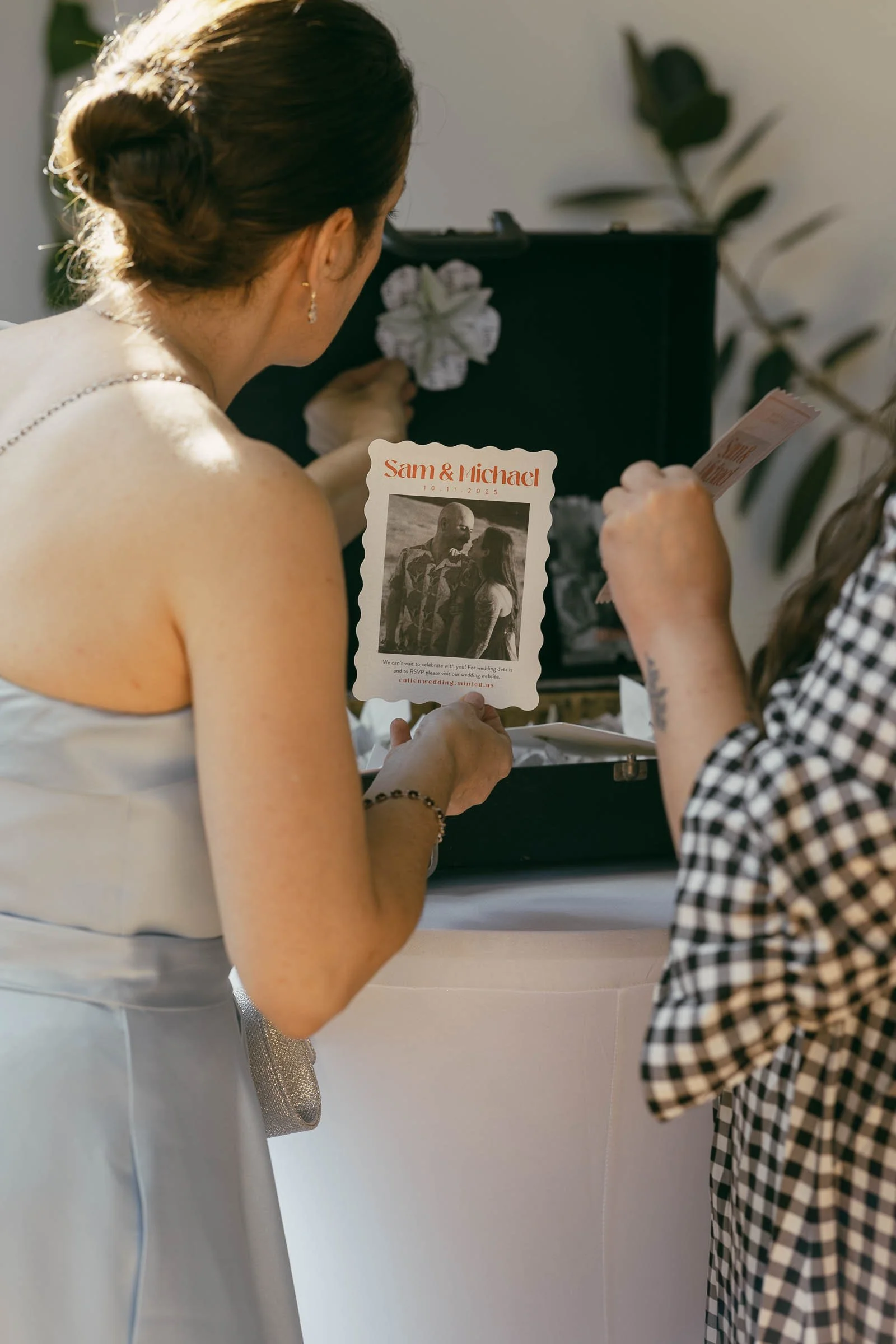  Two women stand at a round table looking into an open suitcase, one holding a vintage-style card with a black-and-white photo and the names "Sam &amp; Michael." The scene is softly lit and decorated with flowers. 