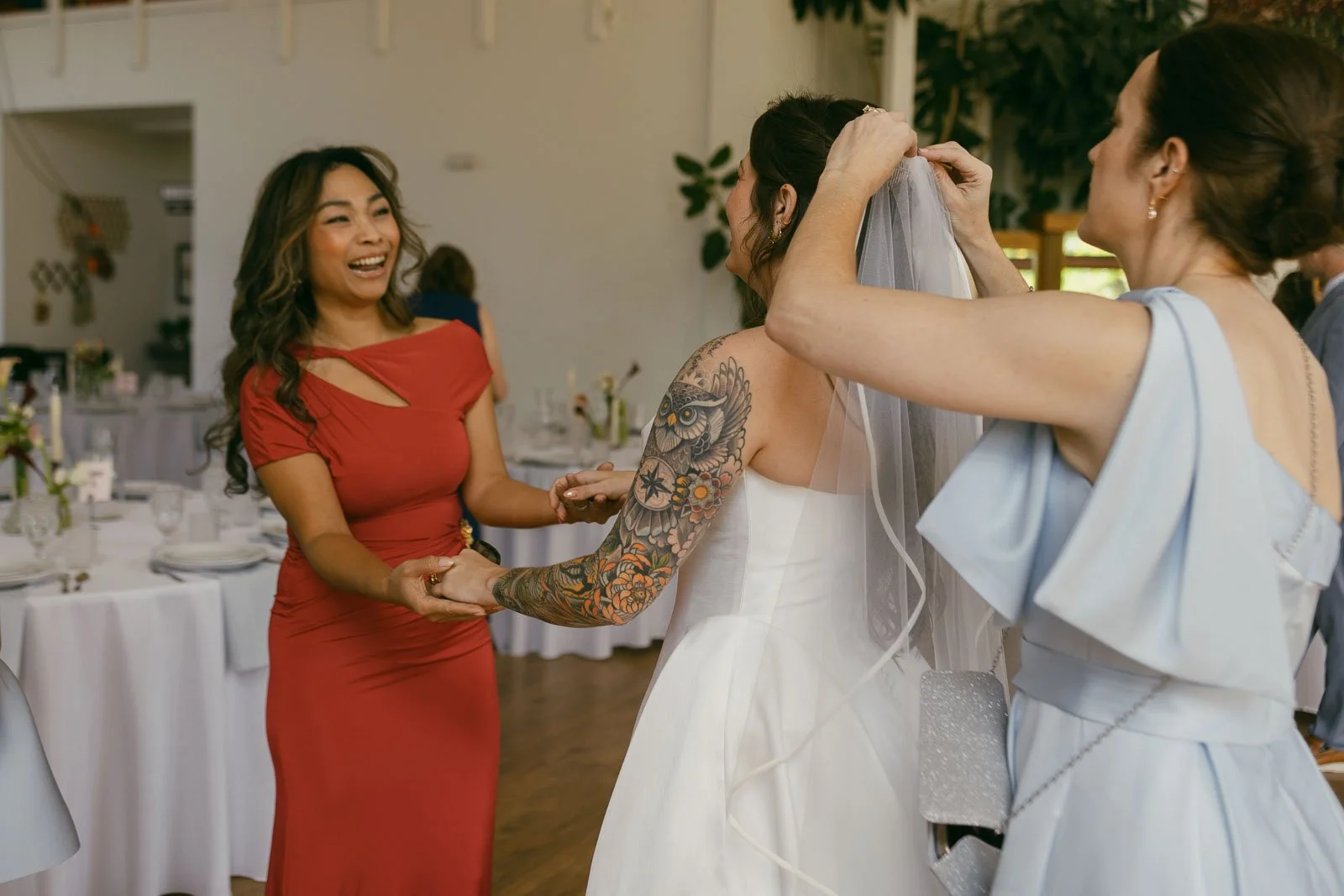  A woman in a red dress smiles and holds hands with a bride in a white gown, while another woman in a light blue dress helps adjust the bride's veil in a decorated indoor venue. 