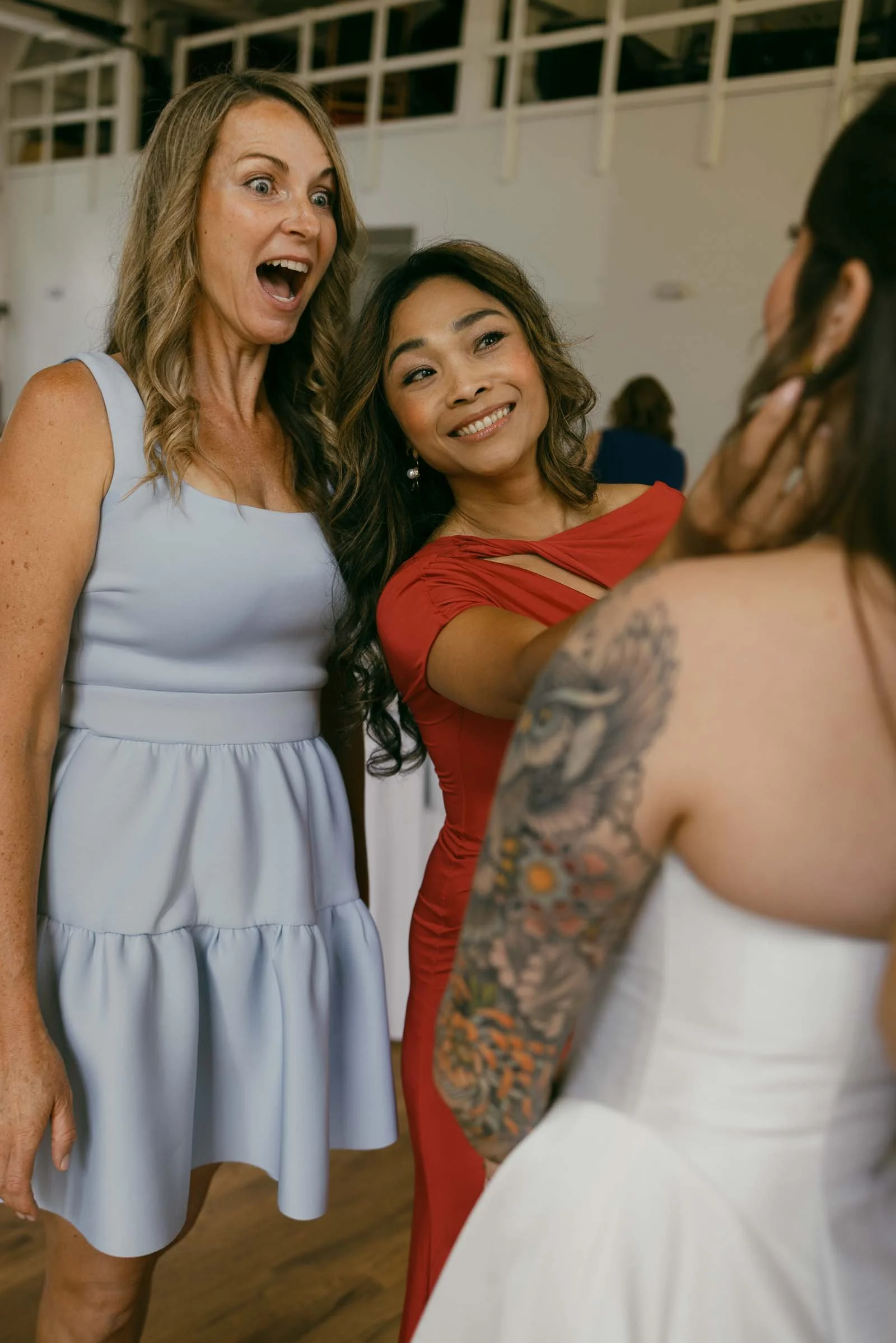  Two women, one in a light blue dress and one in a red dress, interact with a bride in a white gown. The woman in blue looks surprised and happy, while the woman in red smiles warmly. 