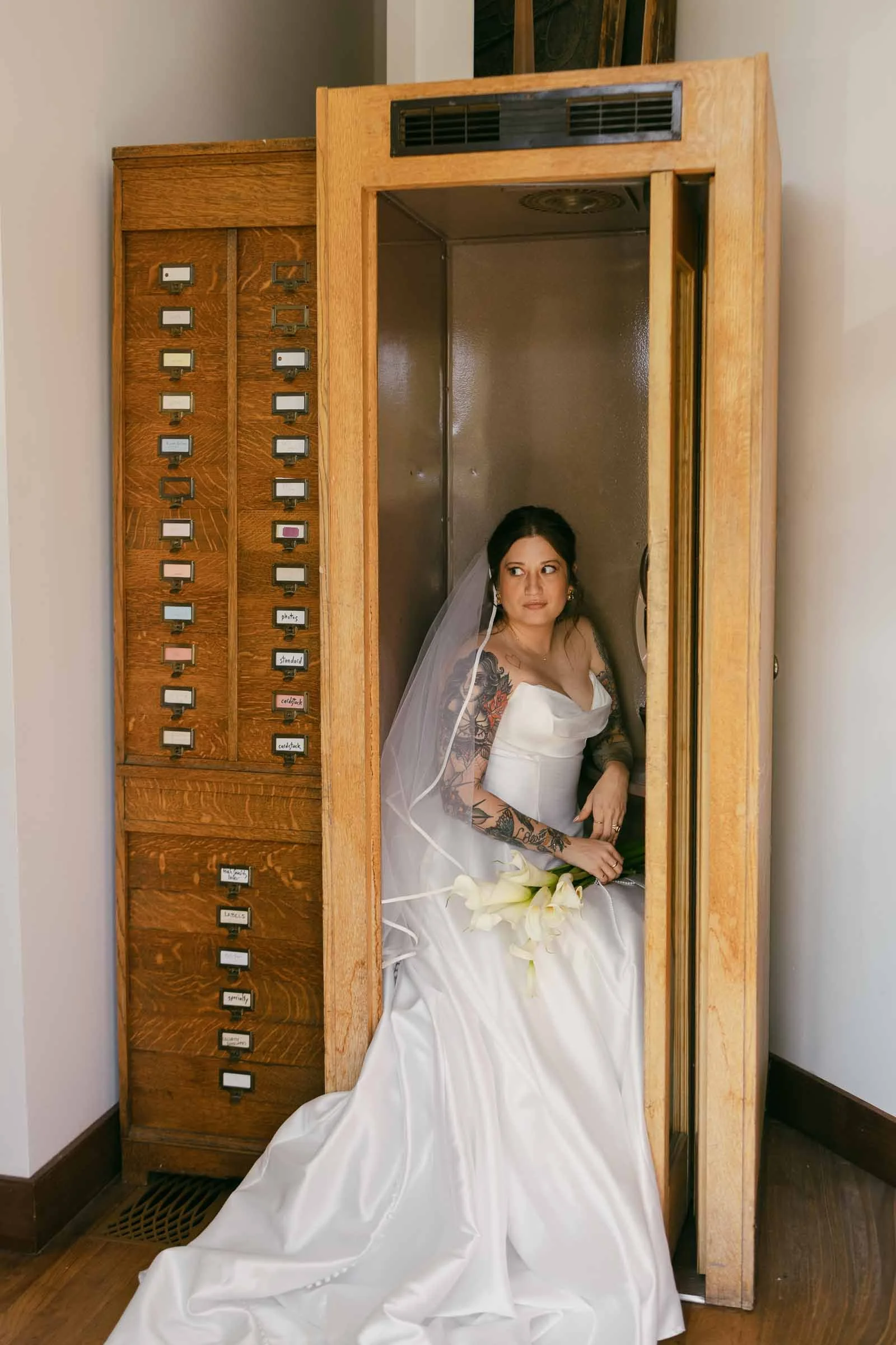  A bride in a white gown and veil sits inside a vintage wooden phone booth, holding a bouquet of white calla lilies and gazing pensively to the side. A wall of labeled wooden drawers is next to the booth. 