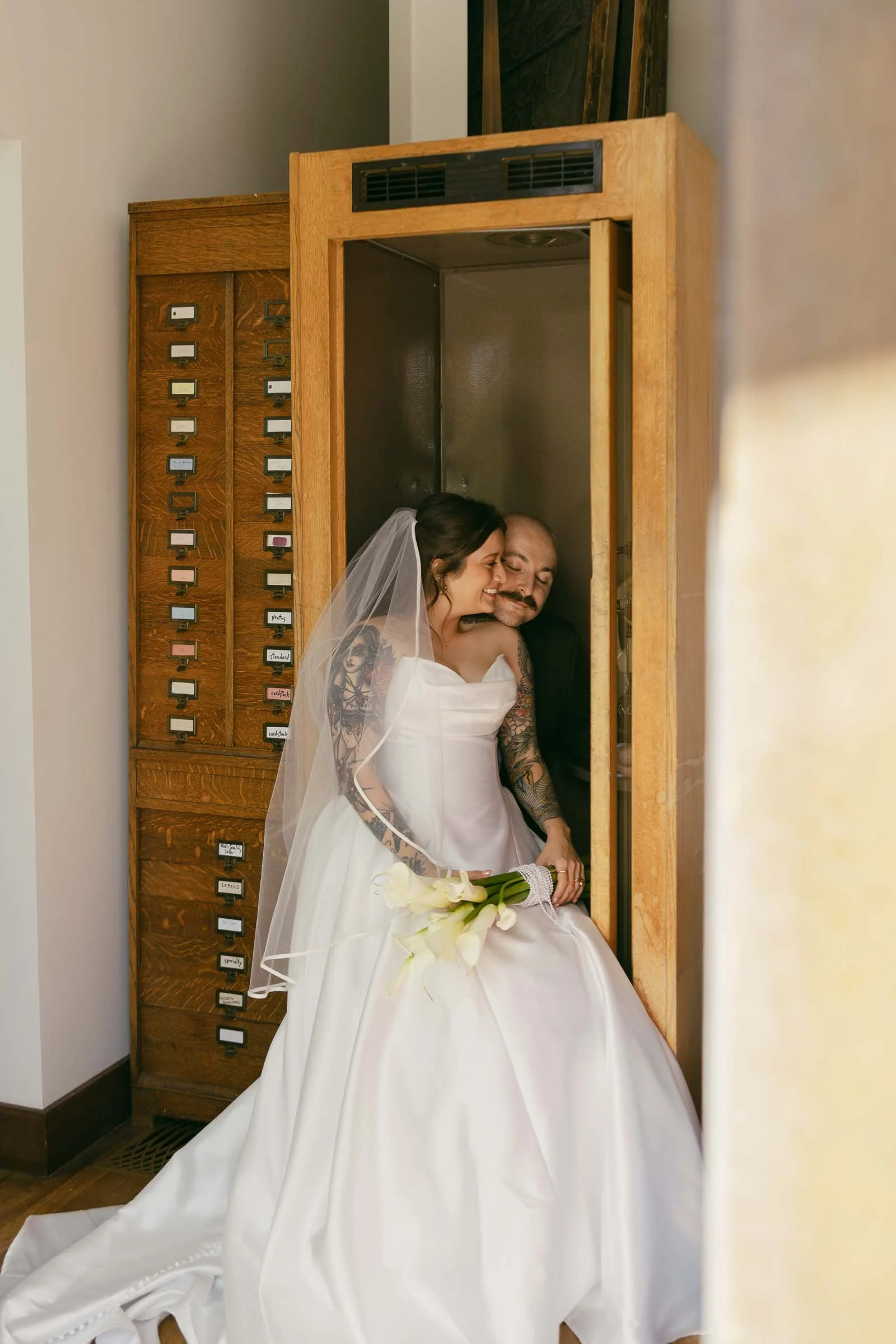  A smiling bride in a white gown and veil sits in a wooden phone booth, holding white lilies, while an older man in a suit lovingly embraces her from behind. A vintage cabinet with small drawers is beside them. 