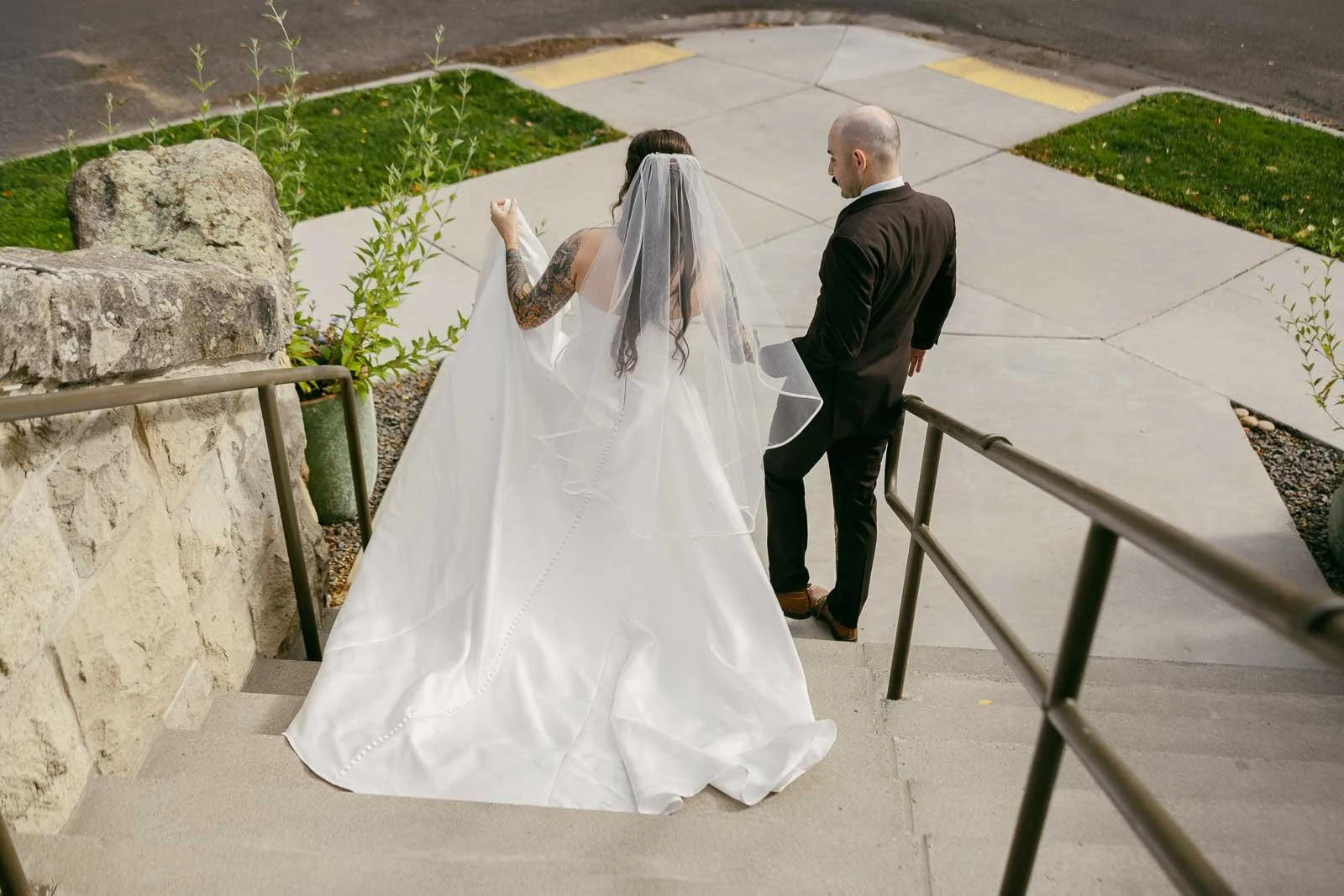  A bride in a white gown and veil walks down outdoor steps with a groom in a dark suit, holding her dress. They are photographed from behind on a sunny day. 