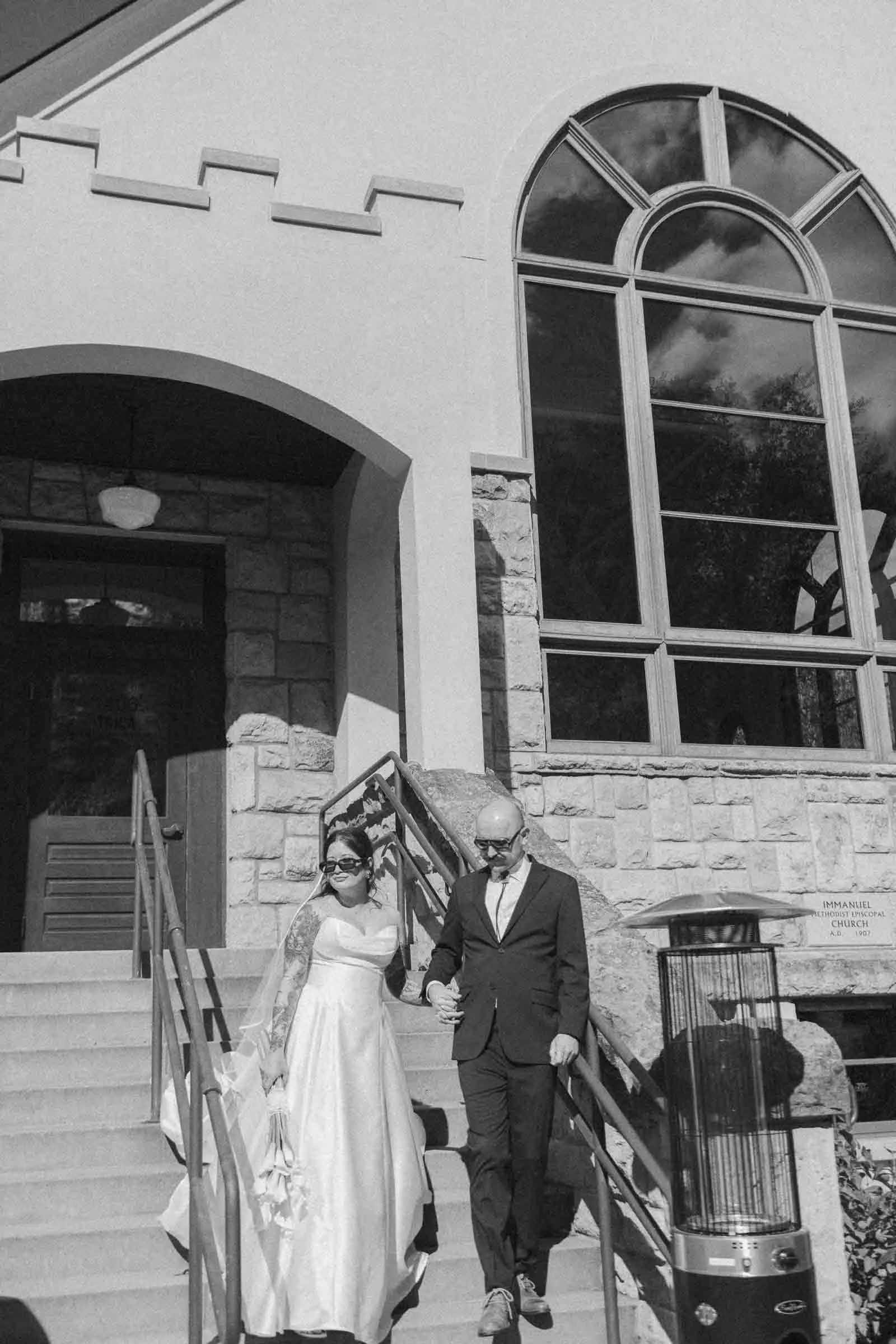  A bride in a strapless gown and veil walks down stone steps with a man in a suit, holding hands outside a building with large arched windows, under bright daylight. 