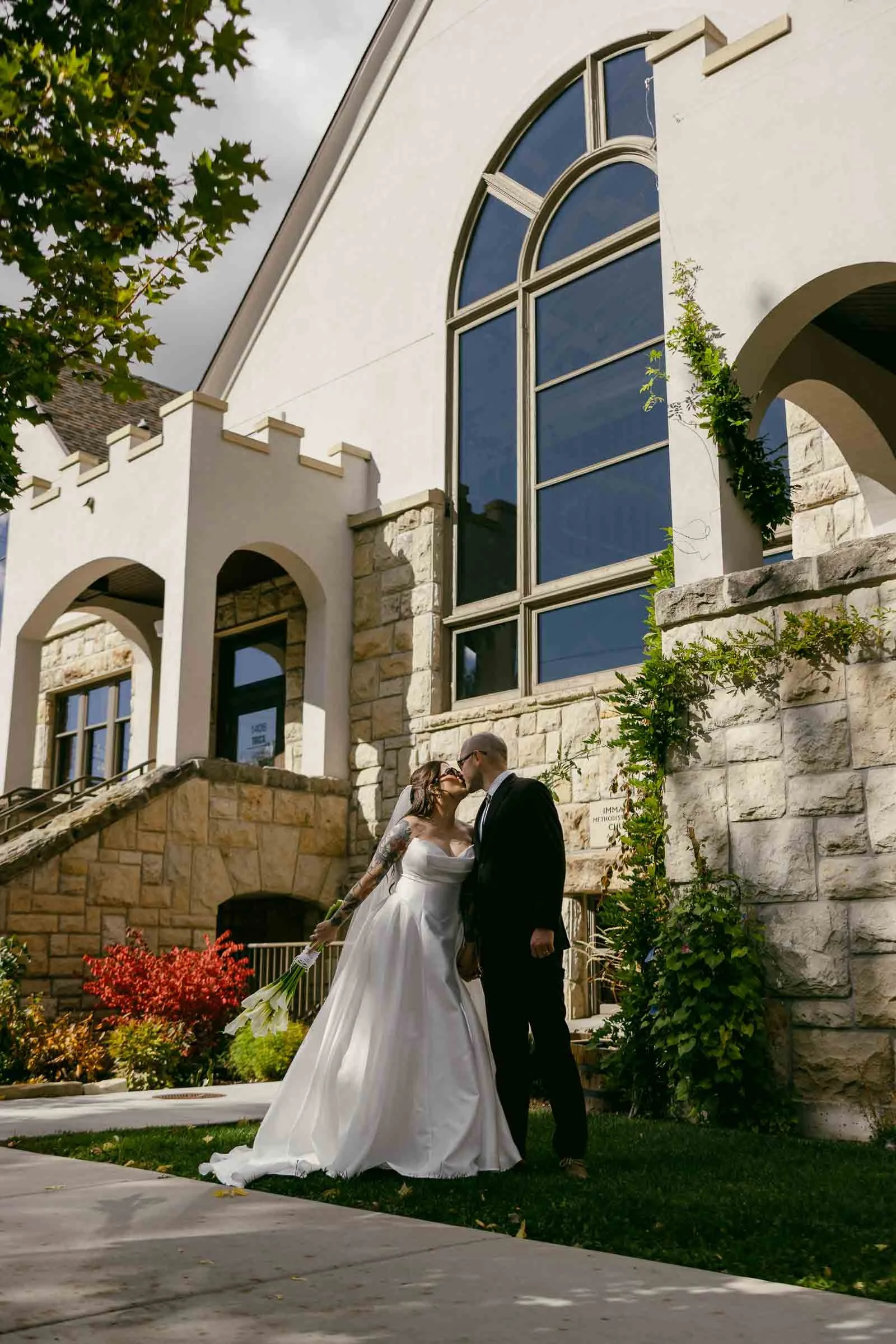  A bride and groom stand outside a stone building with large arched windows, sharing a tender moment. The bride wears a white dress and veil, while the groom wears a black suit. Lush greenery and colorful plants surround them. 