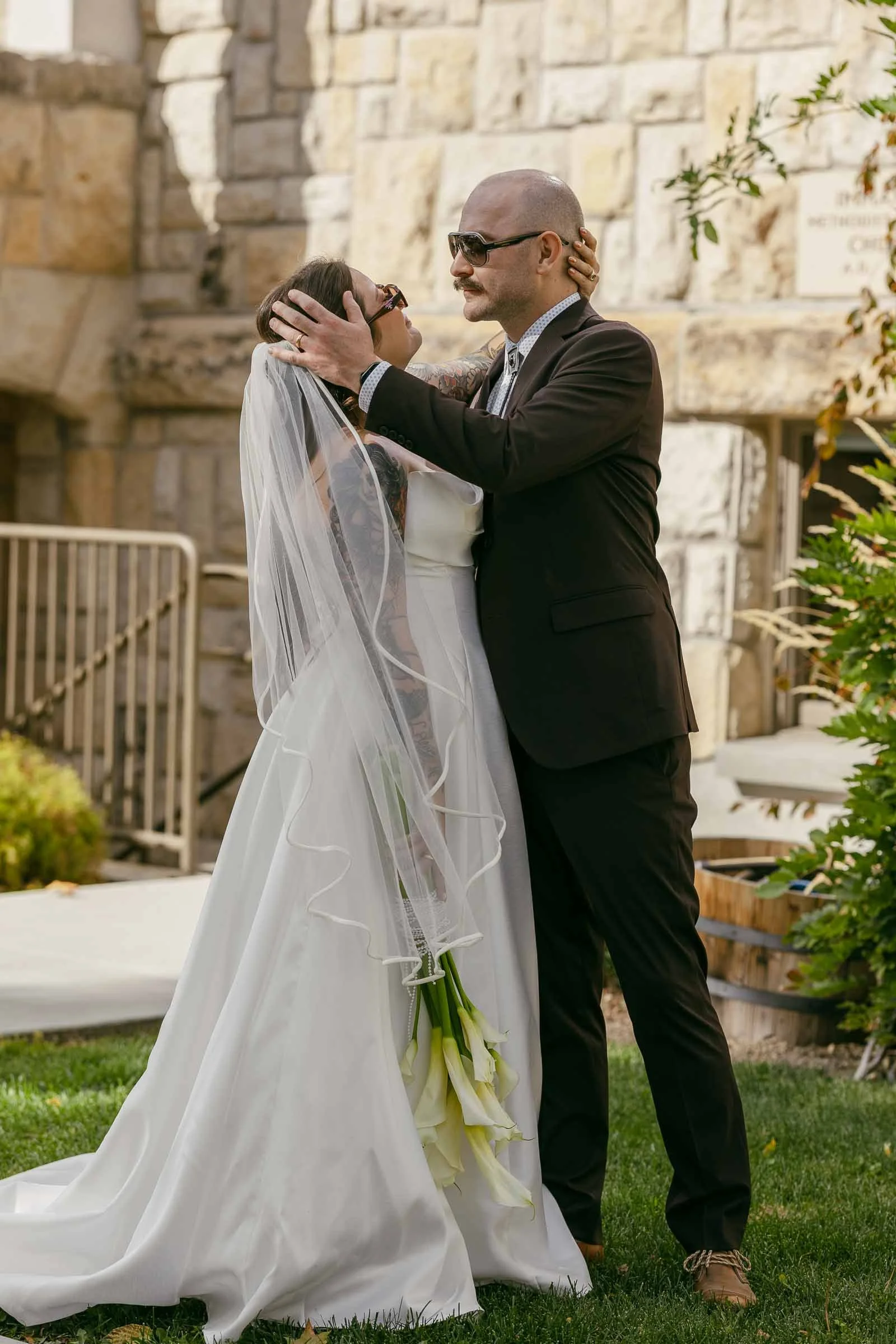  A bride in a white dress and veil holds flowers and embraces a groom in a dark suit and sunglasses. They stand closely, gazing at each other, outside by a stone building and greenery, enjoying a joyful wedding moment. 