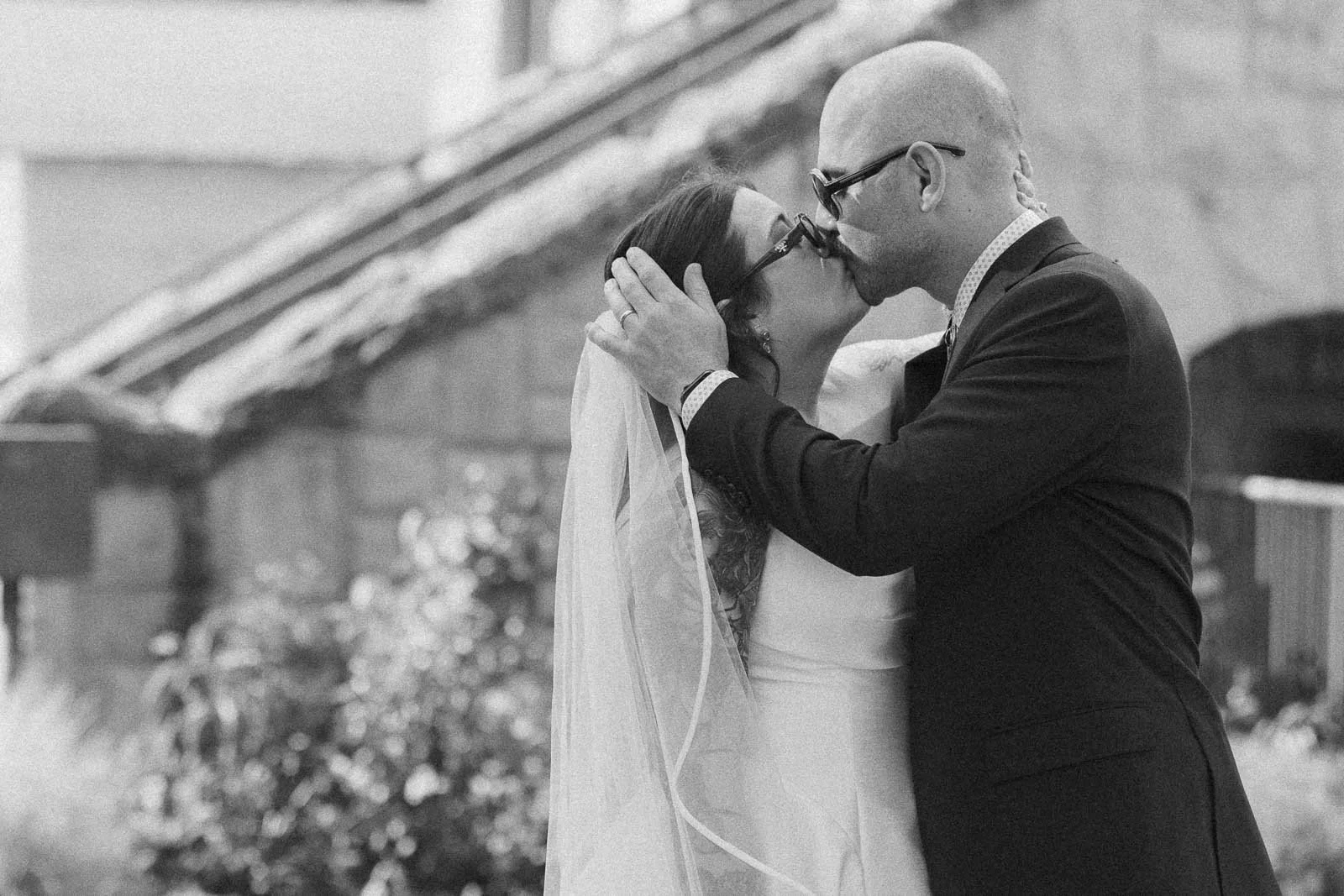  A bride and groom share a kiss outdoors on their wedding day. The bride wears a veil and the groom is in a suit. The image is black and white, with a blurred stone building in the background. 