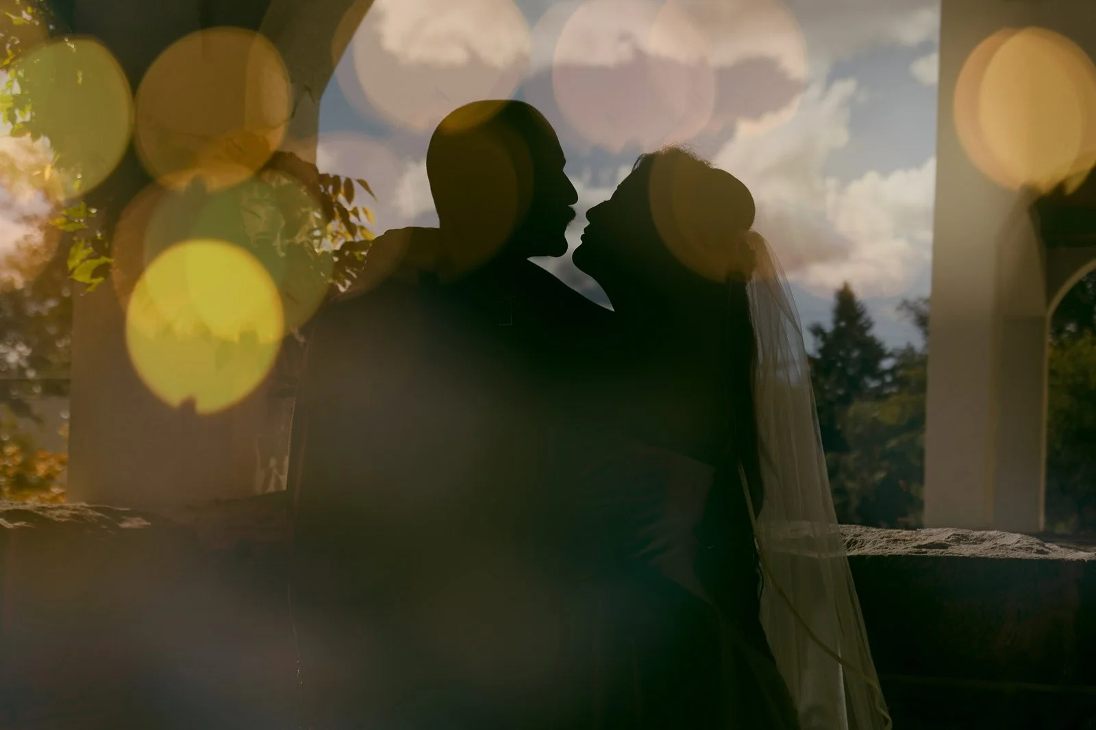  Silhouetted bride and groom face each other closely under an archway, with soft bokeh lights and a cloudy sky in the background, creating a dreamy, romantic atmosphere in this double exposure 