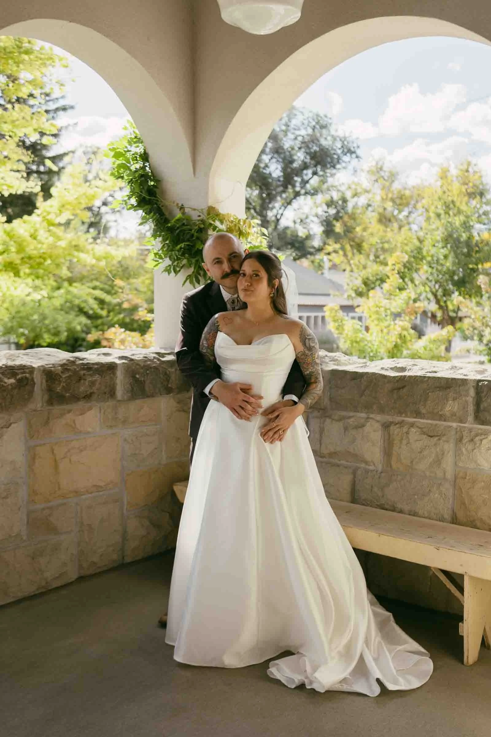  A bride in a white strapless gown stands in front of a groom on a stone porch, surrounded by greenery, as they embrace and look into the distance on a sunny day. 